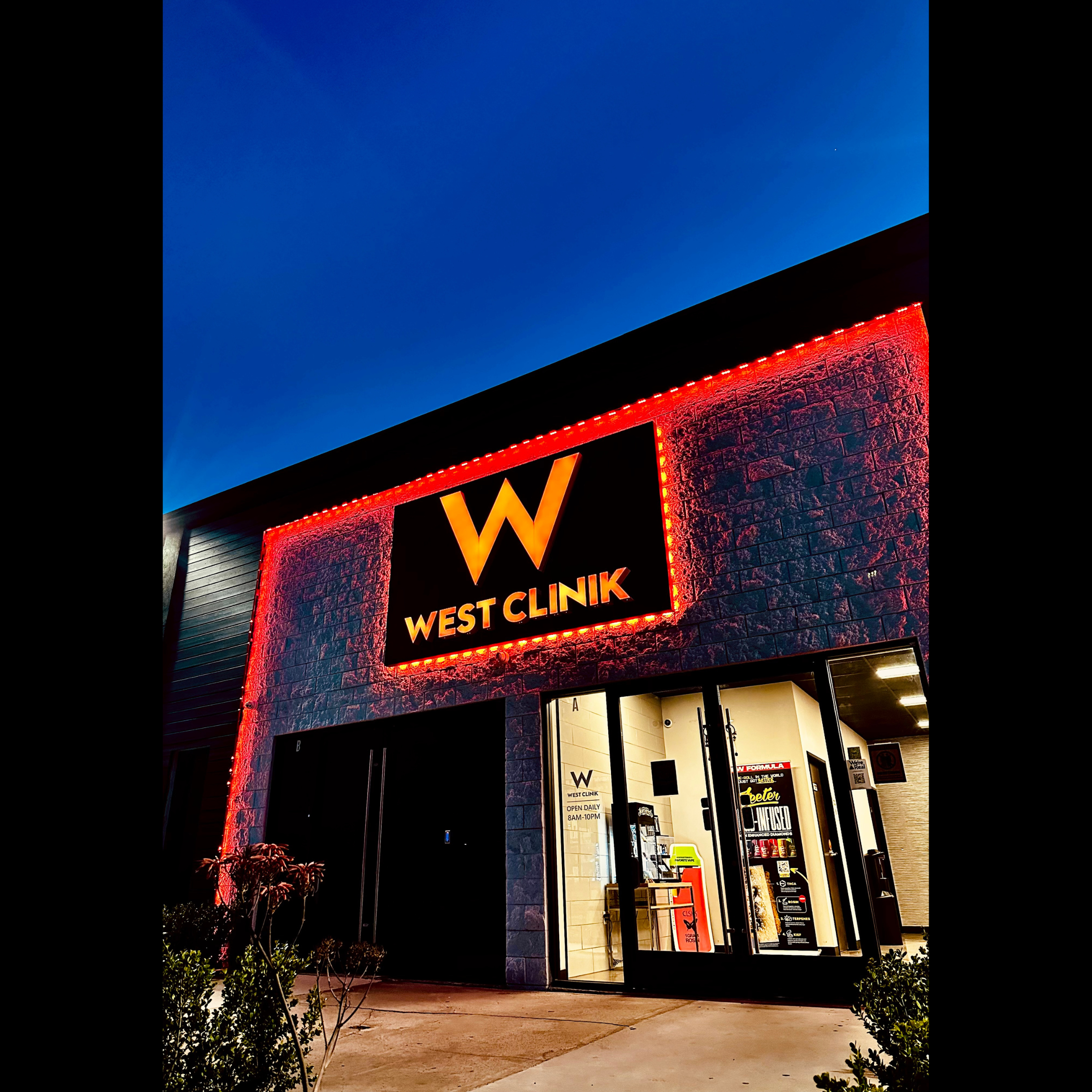 The exterior of a building at dusk with a large illuminated sign reading 'W WEST CLINIK' with a prominent 'W' in the center, decorated with red neon lights outlining the sign and the building's façade. The entrance has glass doors showing the interior with some furniture and promotional posters. Small bushes are planted near the sidewalk in front of the building.