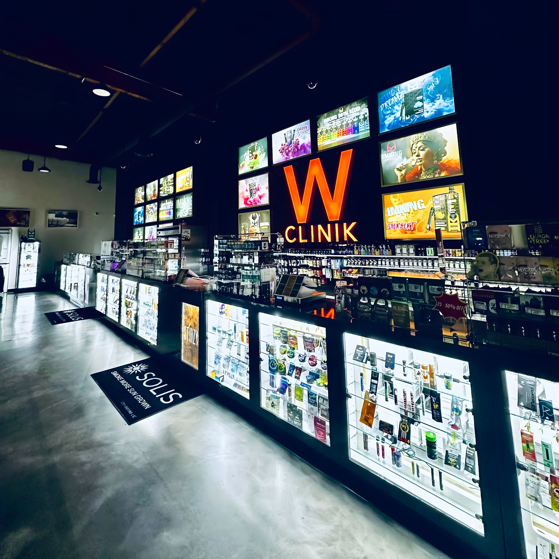 A modern retail store counter with bright backlit display cases containing various health and wellness products, such as vapes, oils, and supplements. A large illuminated sign with a red 'W' and the words 'CLINIK' is displayed on the dark wall behind the counter, accompanied by multiple digital screens showing various advertisements and images.