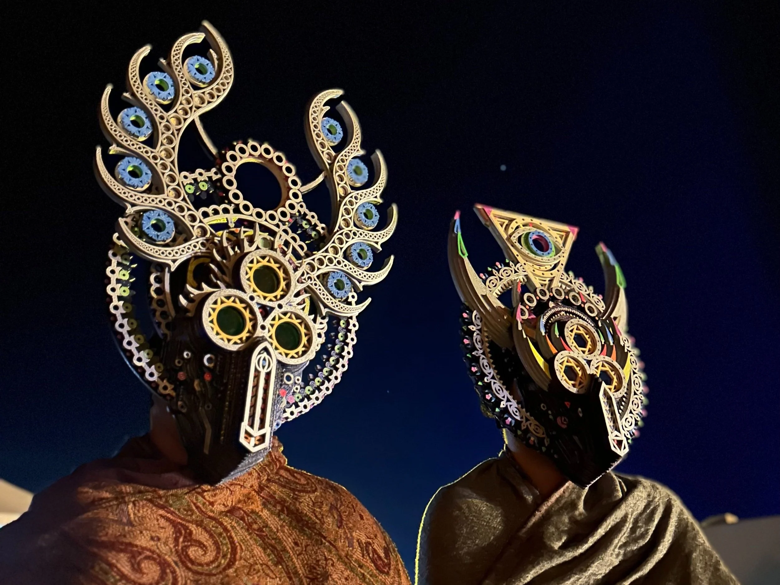 Two people wearing elaborate, colorful, and geometric masks at night against a dark sky.