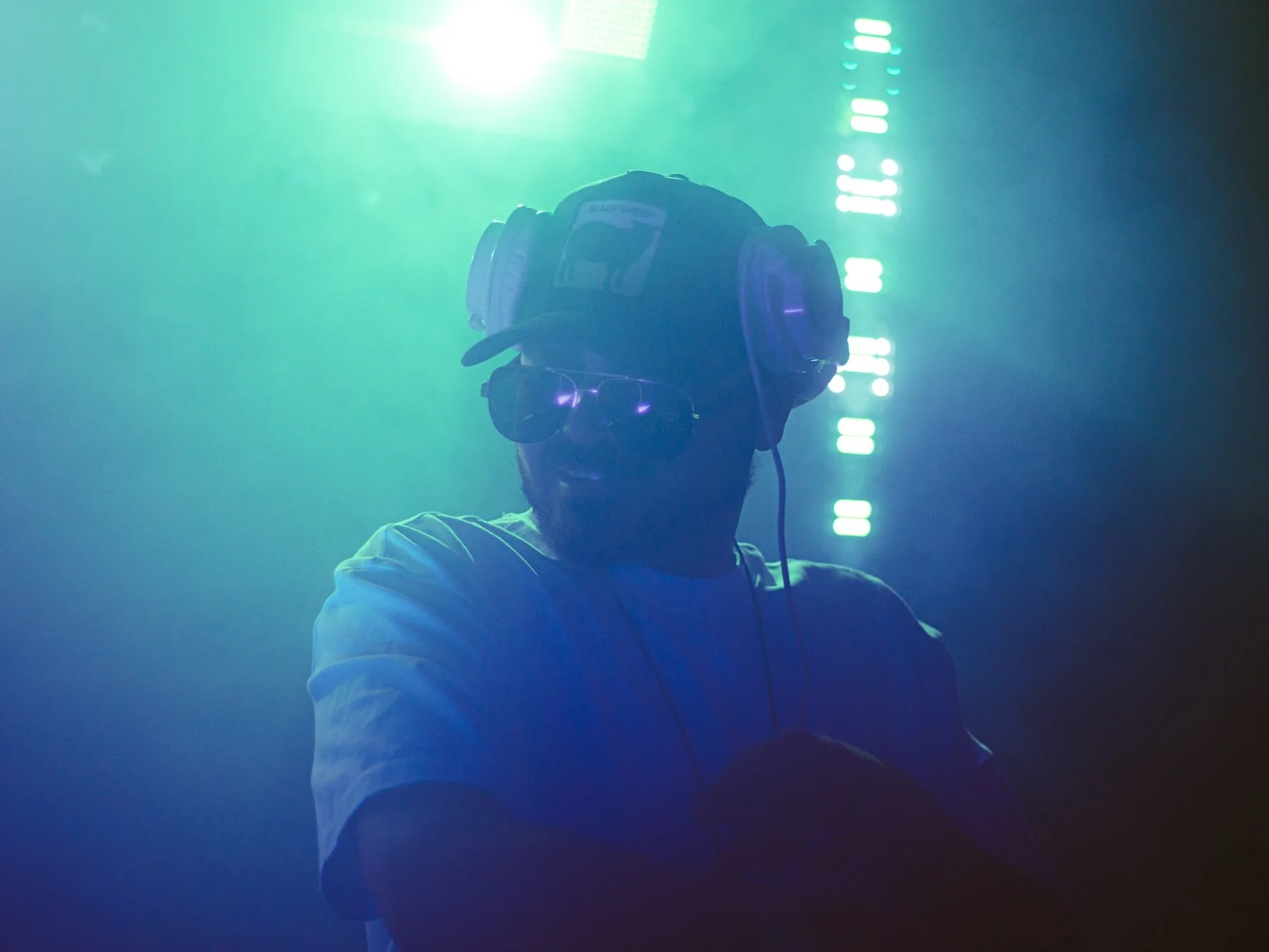 A person wearing sunglasses, a baseball cap, and headphones, in a dimly lit environment with blue and green lighting.