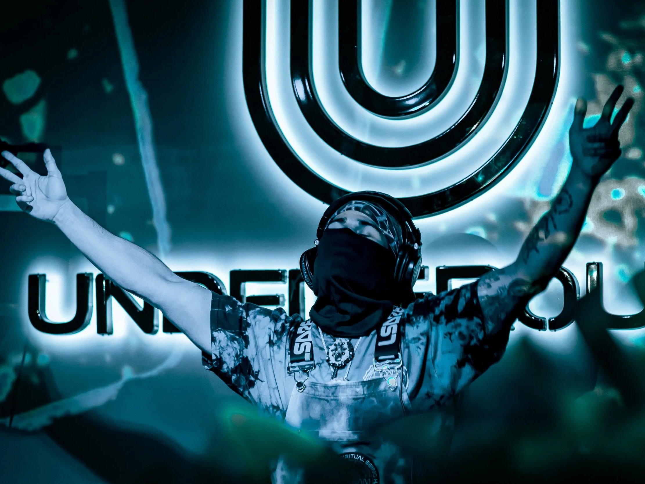 DJ wearing headphones and a mask, raising arms with peace signs in front of illuminated UNEND logo and circular light installation.