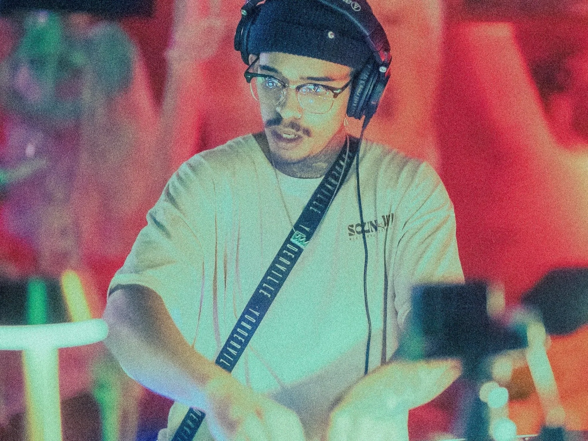 A young man with glasses, wearing a beige shirt and headphones, standing at a DJ booth with colorful lighting in the background.