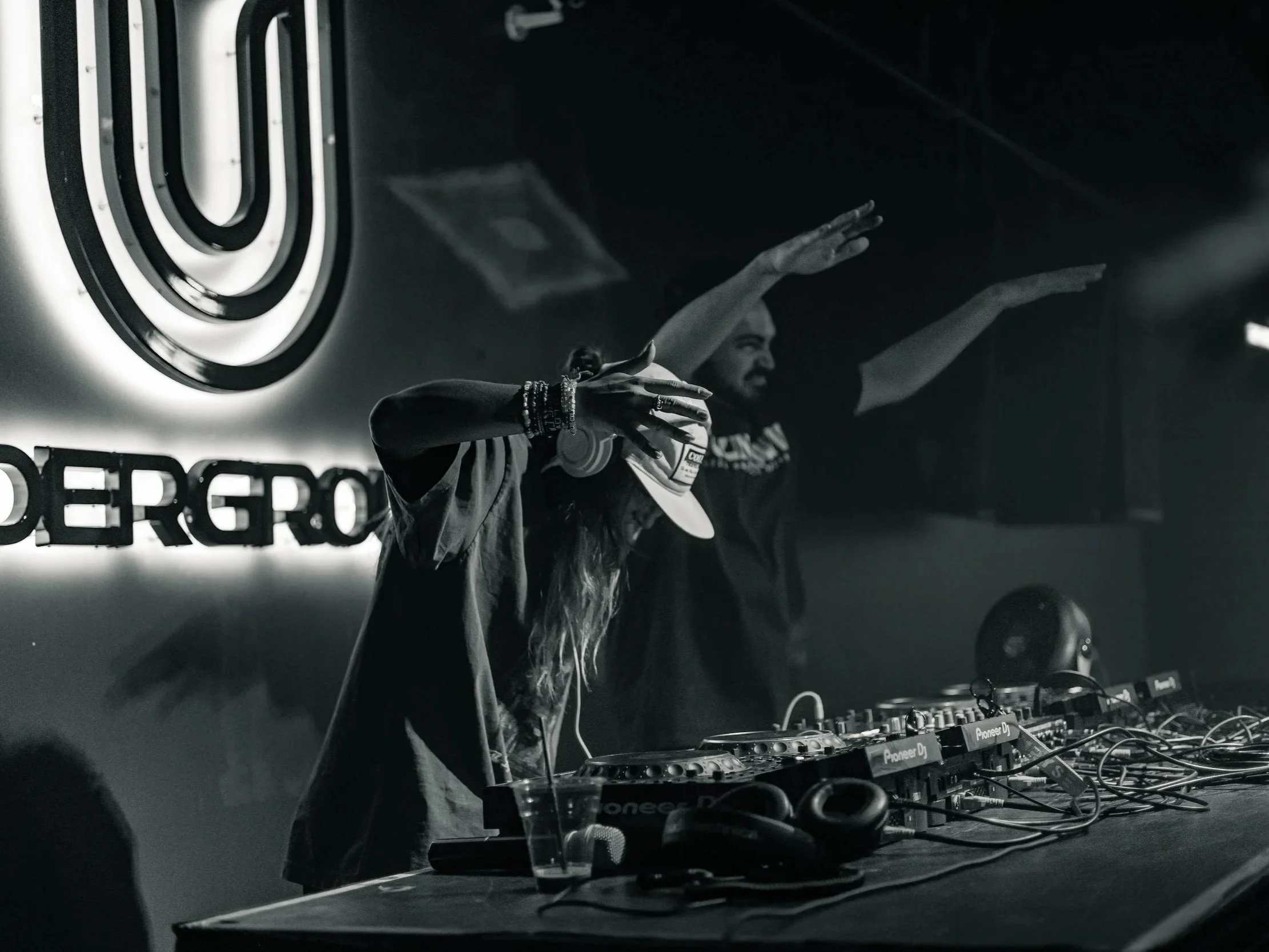 A DJ performs behind a table with mixers and turntables at a nightclub, with a person dancing in front and a neon sign reading 'UNDERGROUND' in the background.