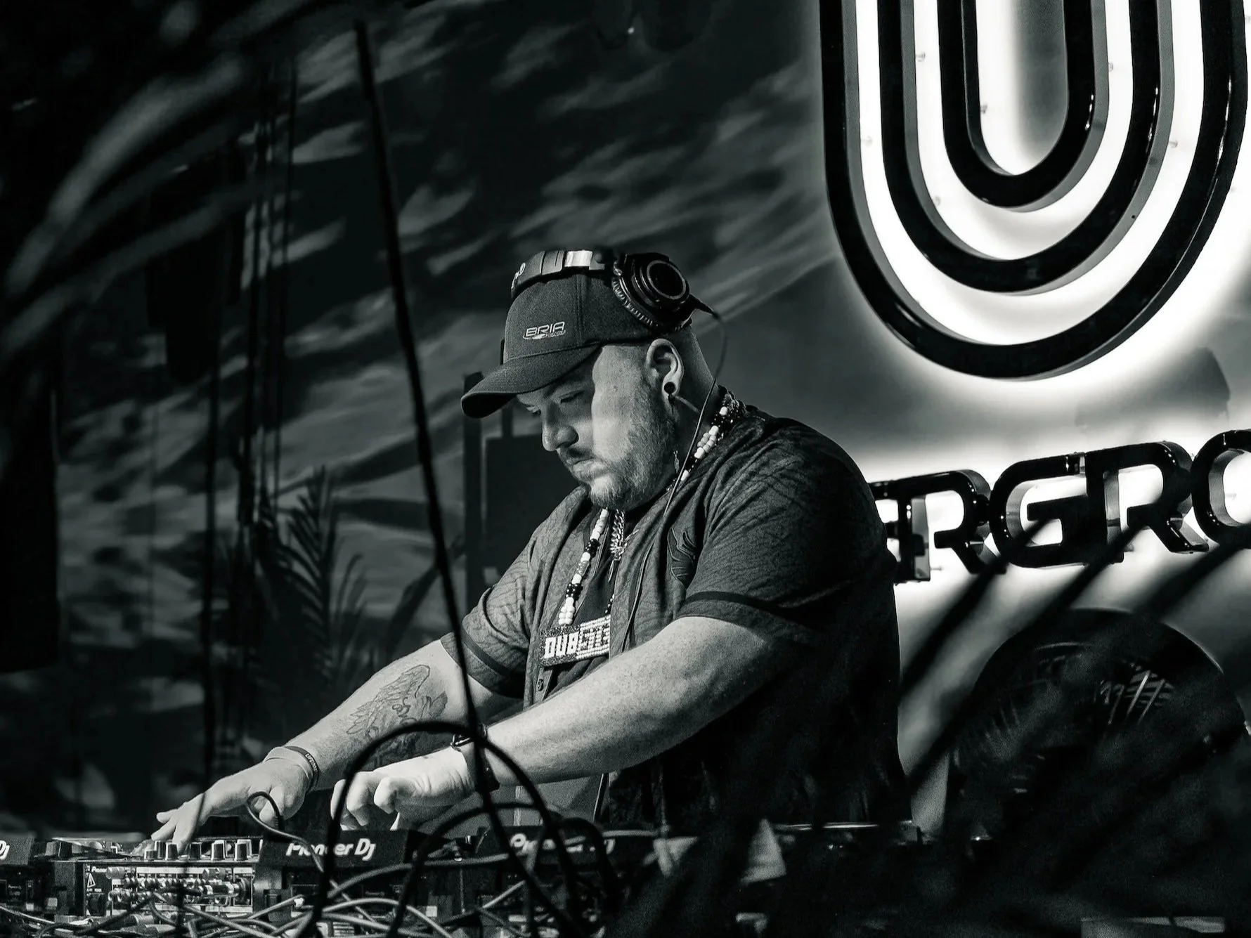 A DJ wearing a cap and headphones, working at a mixer in a dimly lit venue with a large illuminated target symbol and partial text in the background.