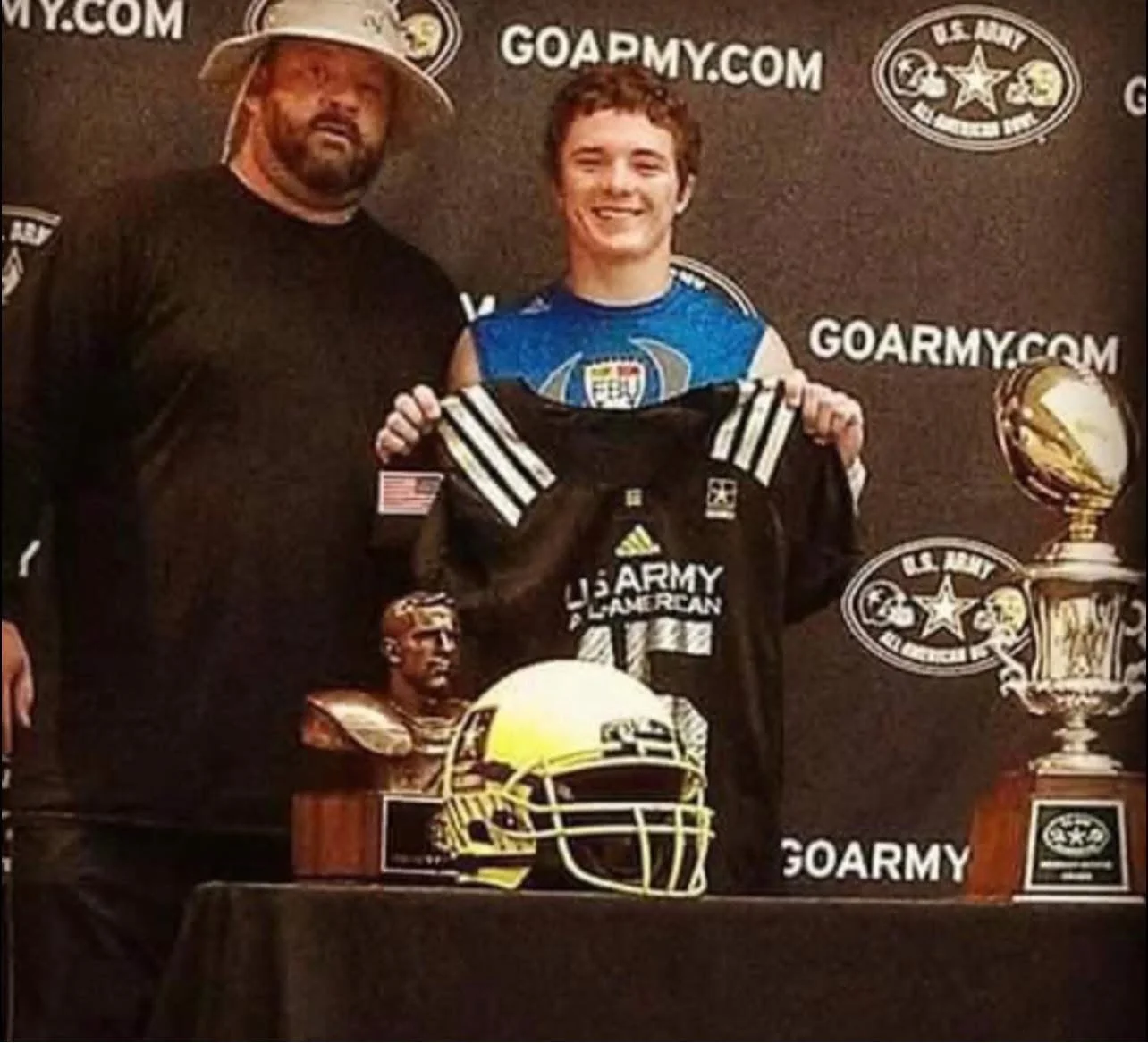 Tex Kelly smiling while holding up a black football jersey during a ceremony, standing beside his father, who wears a beard and wide-brimmed hat. The background features GOARMY.COM