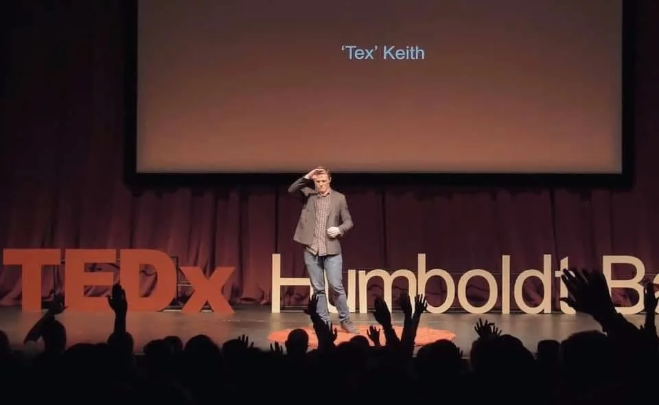 Tex Kelly giving a TEDx talk on stage at TEDxHumboldt, standing with one hand on his head as audience members raise their hands—captured during a live speaking event in Humboldt County, California. Tex Kelly is a creative director and videographer.