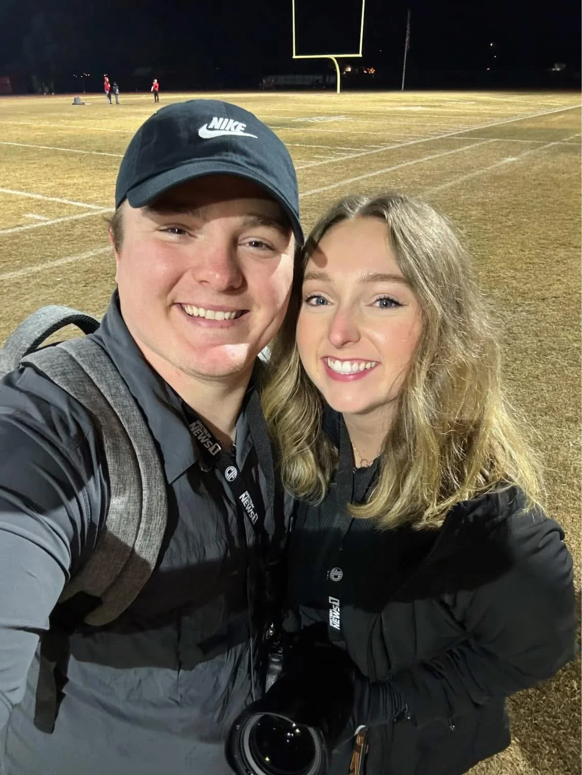 Tex Kelly and his wife, Molly, smiling and taking a selfie on a lit football field at night in Humboldt County, California. Tex is wearing a black Nike cap and camera gear, while Molly has long blonde hair and a black jacket. 