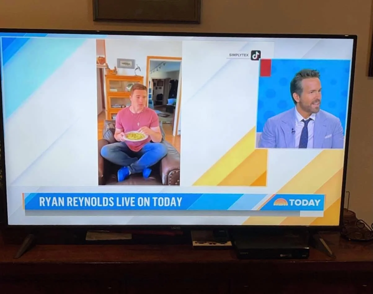 Television screen showing a split image of Tex Kelly sitting on a couch eating food at home, and actor Ryan Reynolds speaking on a news program with the “Today” show logo and the text ‘Ryan Reynolds live on Today’—a humorous juxtaposition.