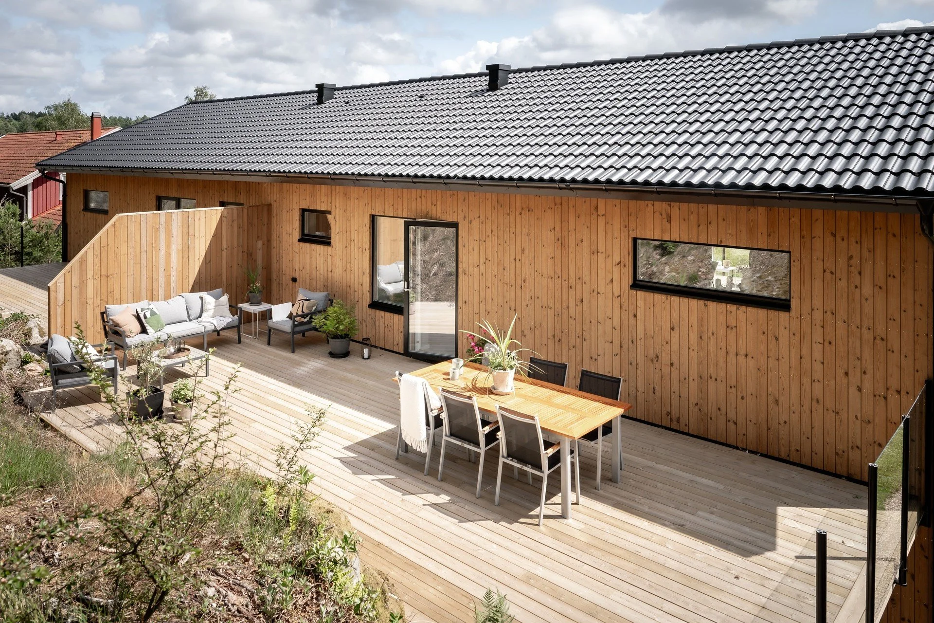 Trädäckad uteplats med möbler och växter framför ett hus med träfasad och svart tak.