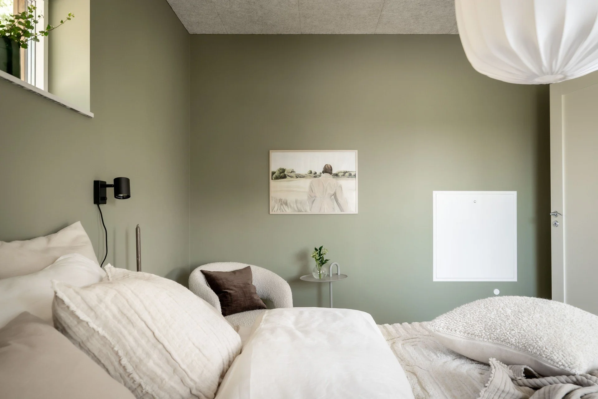 Sovrum med gröna väggar, vita sängkläder, en sittmöbel med kuddar, växter, en konstposter och en taklampa.