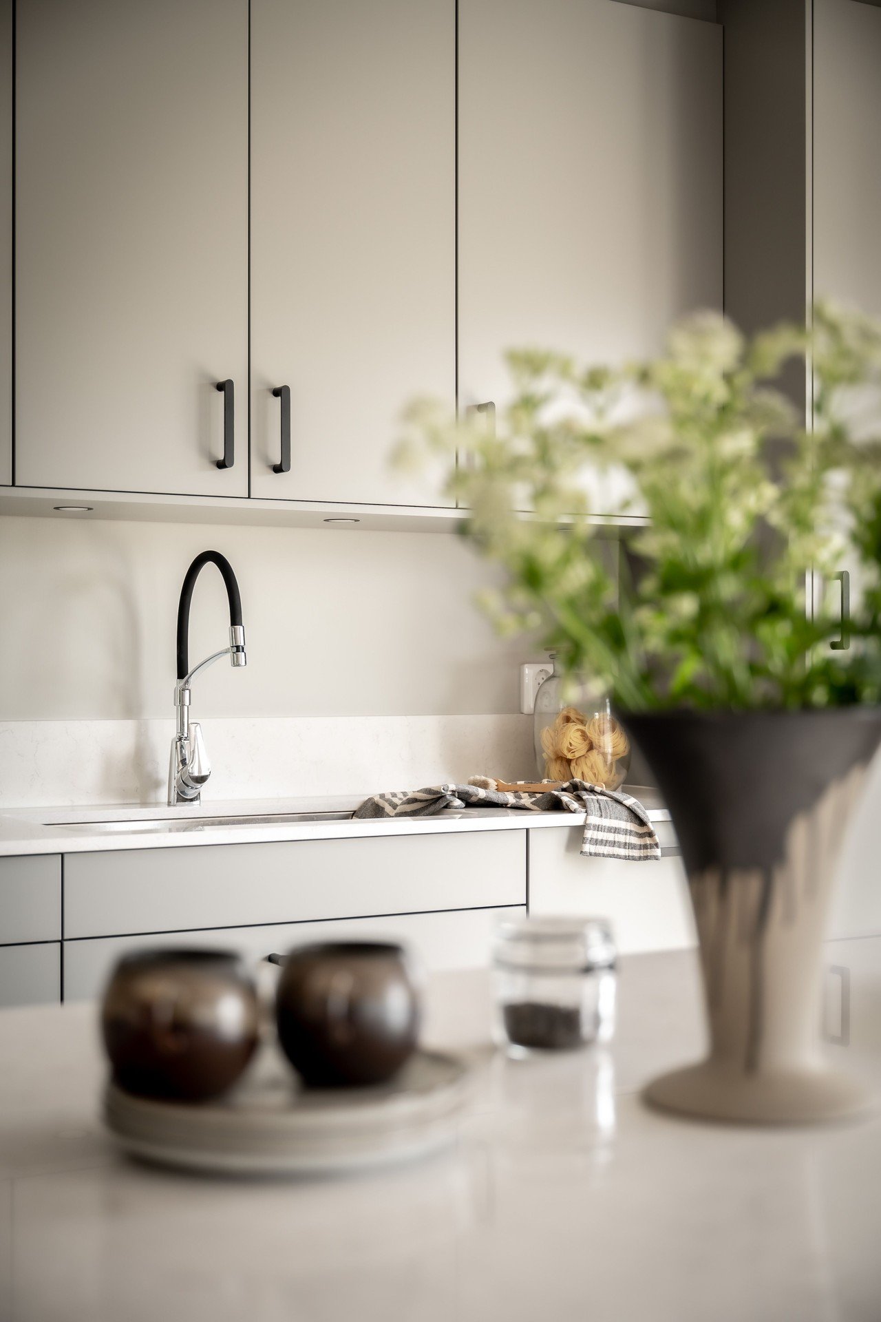 Modern köksinredning med ljusgrå skåp, svart handtag, runt vitt kaklat diskbänk, svart kökskran, blommor i en vas, några bakverk på en tallrik, köksredskap och köpmanöver i bakgrunden.