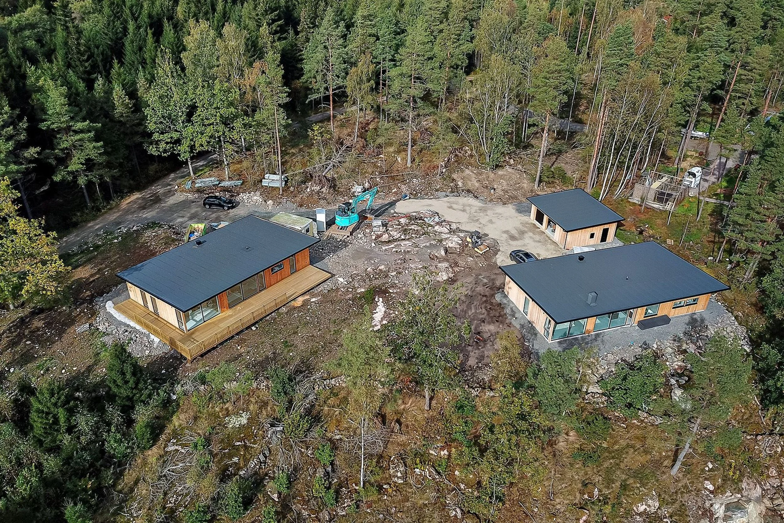 Luftbild av tre små moderna hus i skogen, två större och ett mindre. Omkringliggande skog och en grävmaskin synlig. Höstlandskap.