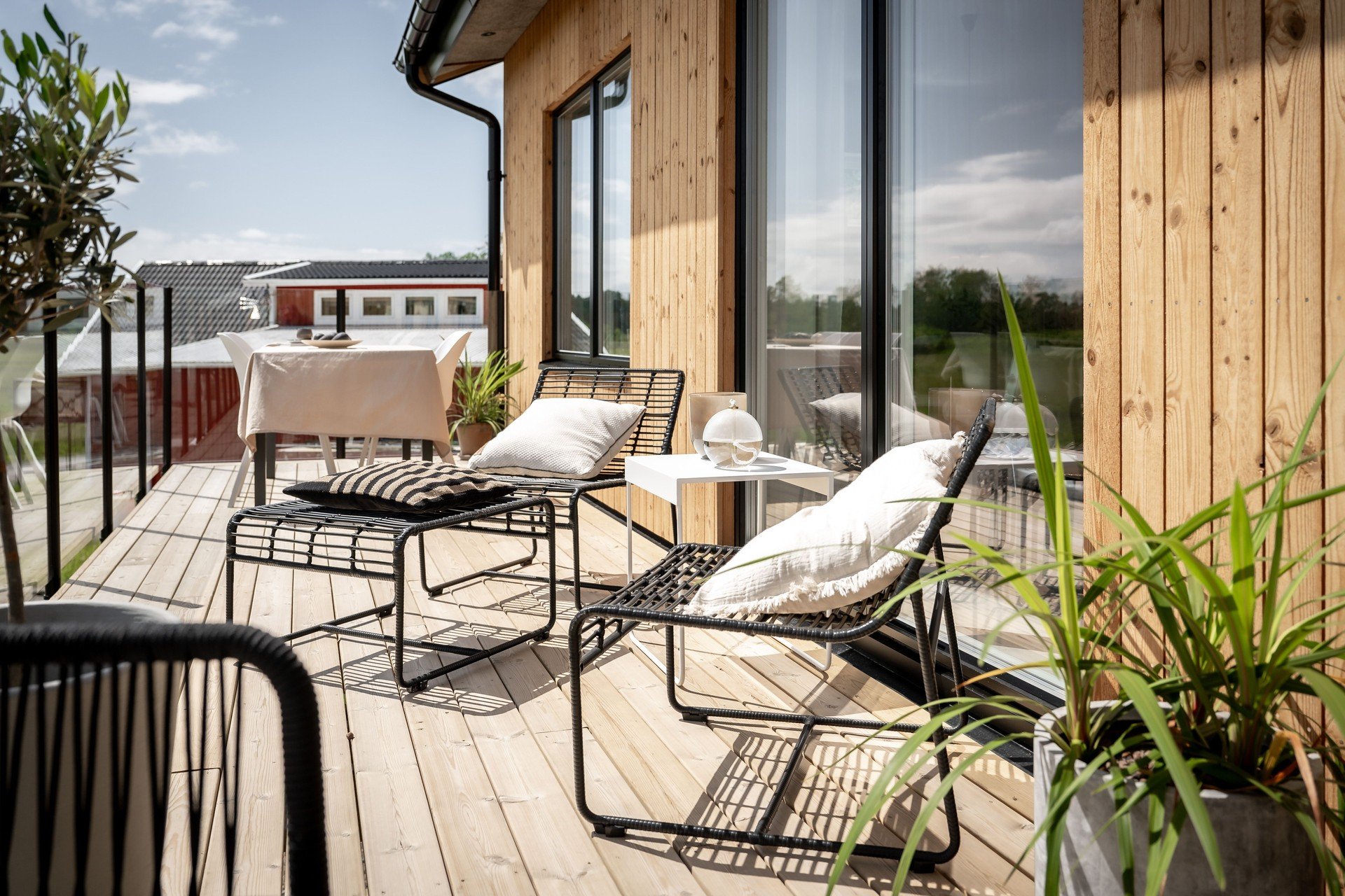 Träveranda med moderna metallstolar, kuddar och växter, bord med duk, stora fönster, soligt väder.