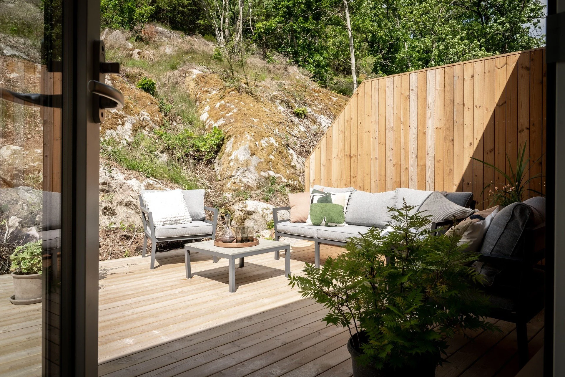 Uteplats med sittmöbler avgränsad av en tallskog och en trädspärr i trä, vilande på en trädäckad veranda, med växter i krukor och ett glasvas.