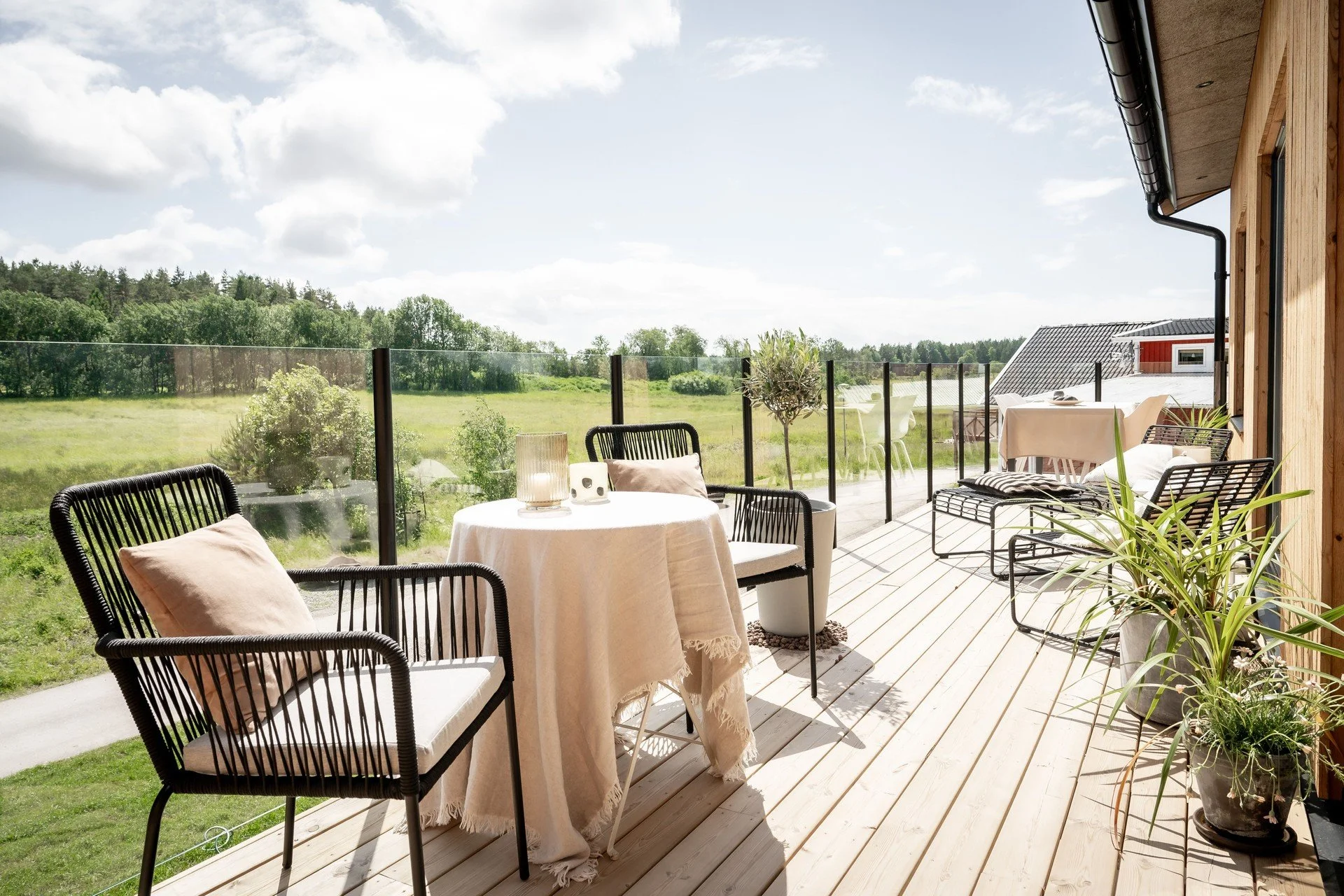 Solig terrass med träplank, stilrena utemöbler, glasräcke och utsikt över en grönskande äng. Bord med duk och kuddar, samt krukväxter som dekoration. Ljusa och luftiga känsla med mycket dagsljus.