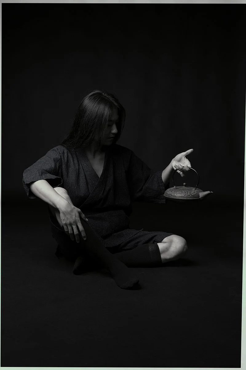 A black and white photo of a person sitting cross-legged, wearing dark traditional clothing, holding a decorative teapot in a dimly lit setting.