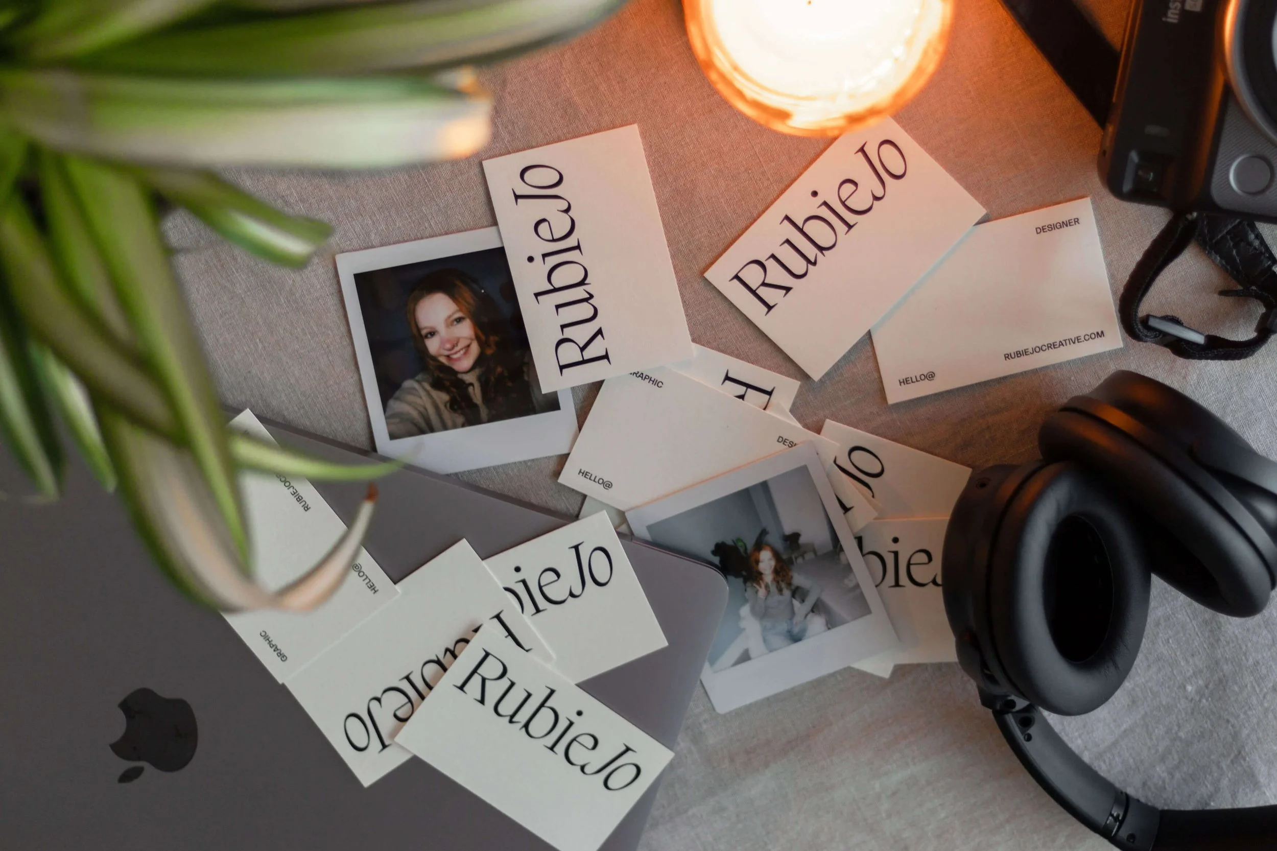 A desk with business cards, printed photos, a candle, headphones, and a camera, with a partial view of a green plant and a logo on the desk.