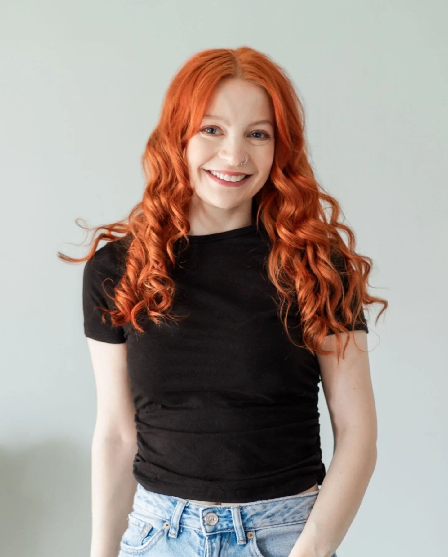 A derby freelance designer with long, curly red hair, smiling, wearing a black T-shirt and light blue jeans, standing against a plain, light-coloured background.