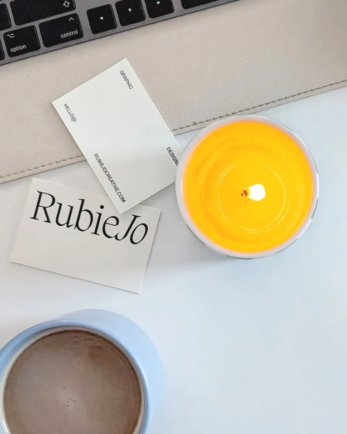 Derby graphic designer Rubie Jo Creative's business card design lay on the desk with an overhead view of a candle, cup of coffee and keyboard on the white desk.