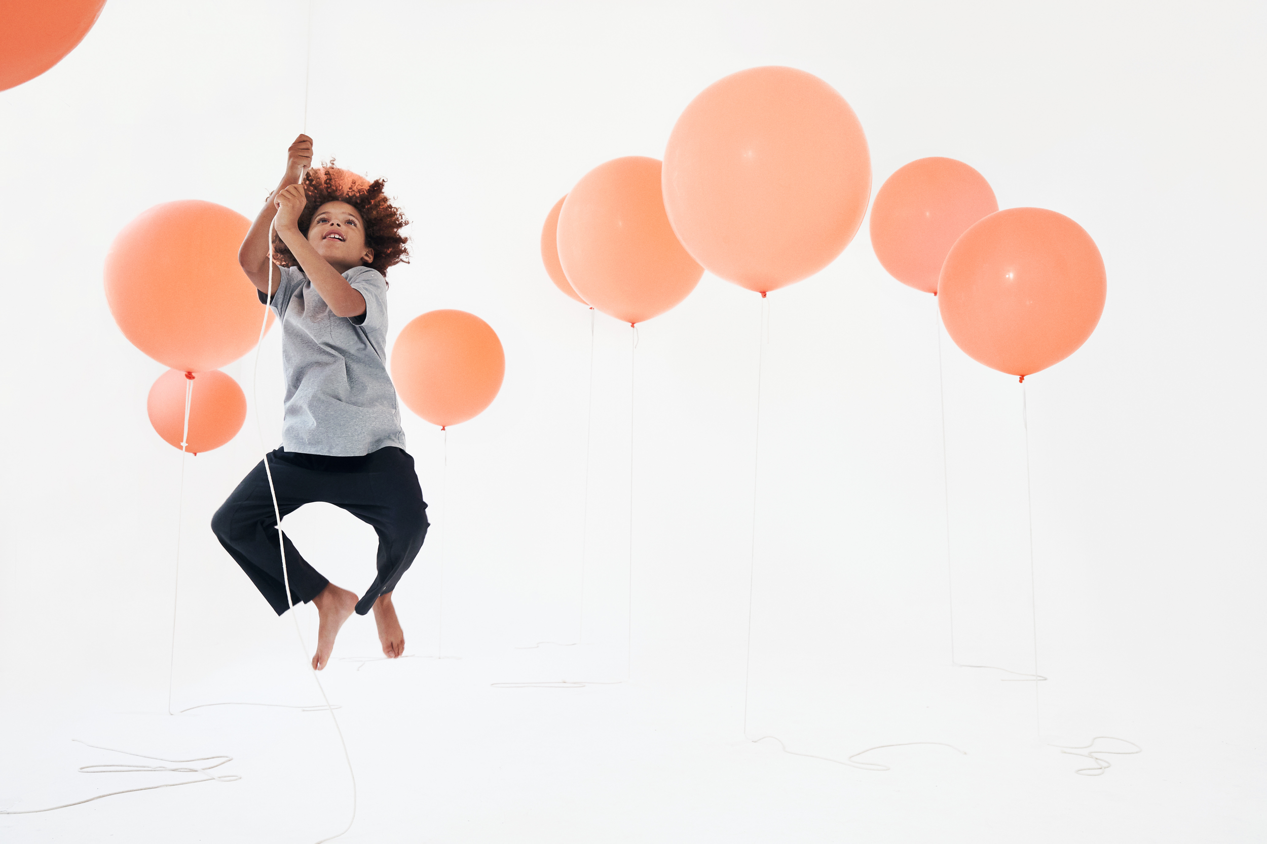 A young girl with curly hair floating in the air holding onto a string connected to a peach-colored balloon, surrounded by several other peach-colored balloons on a white background.