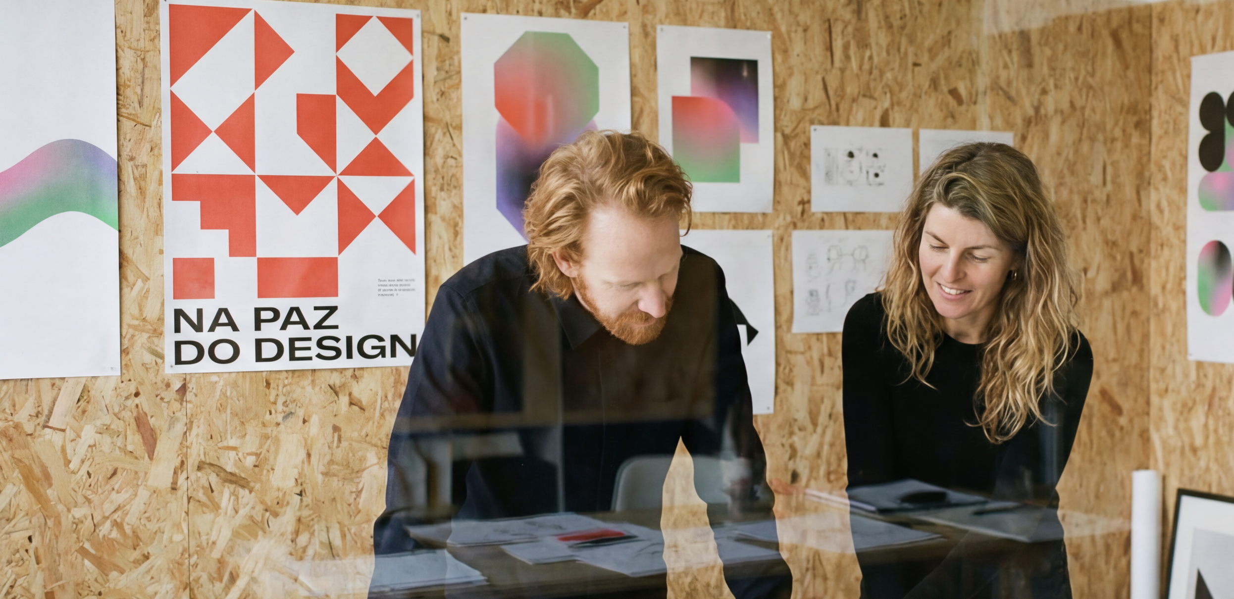 Two people, a man with red hair and a woman with blond curly hair, are smiling and discussing something in a design studio. They are standing behind a glass window, with various colorful abstract posters and sketches on a wooden wall in the background.