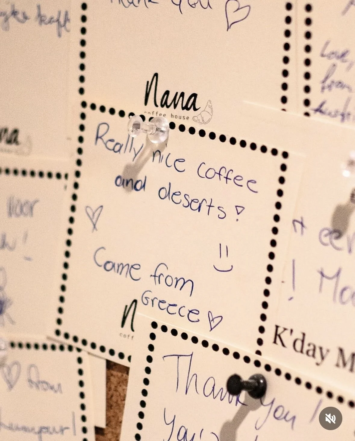 Handwritten note on a yellow sticky note stating, 'Really nice coffee and desserts! Came from Greece,' with small hearts and smiley faces, pinned to a corkboard with push pins.