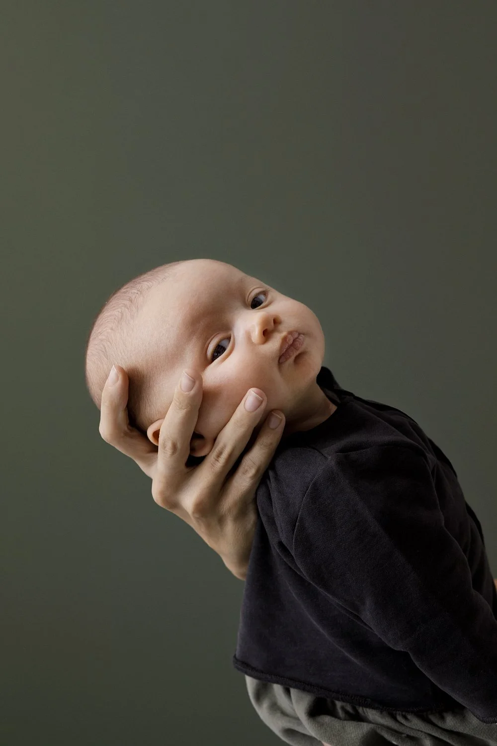 A close-up of a young child with a shaved head, resting their face in a hand, with an adult's hand supporting their head against a plain, muted green background.