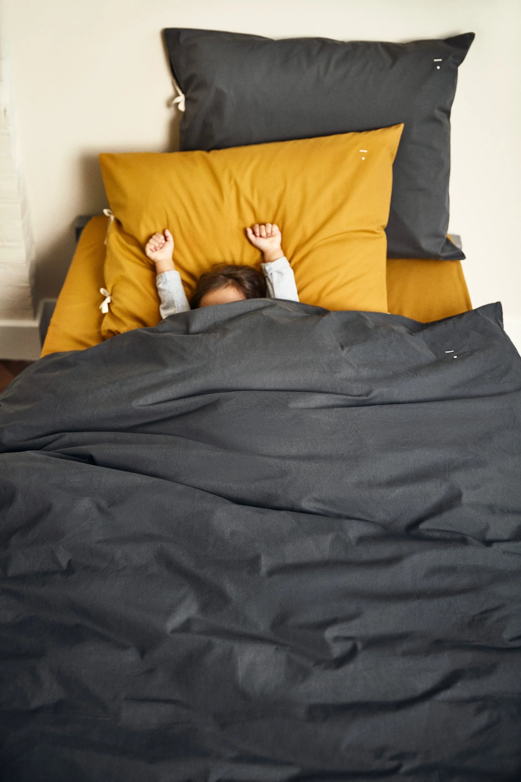 A child lying in bed with arms raised, covered with dark gray and mustard yellow bedding, and matching pillows.