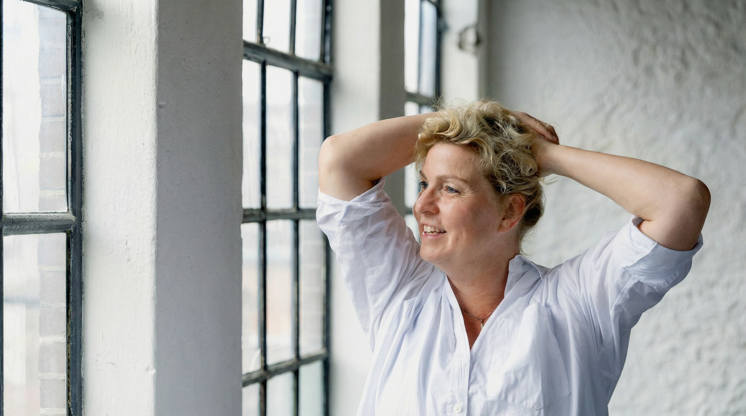 A woman smiling with her hands behind her head, standing near a large industrial-style window in a bright room.