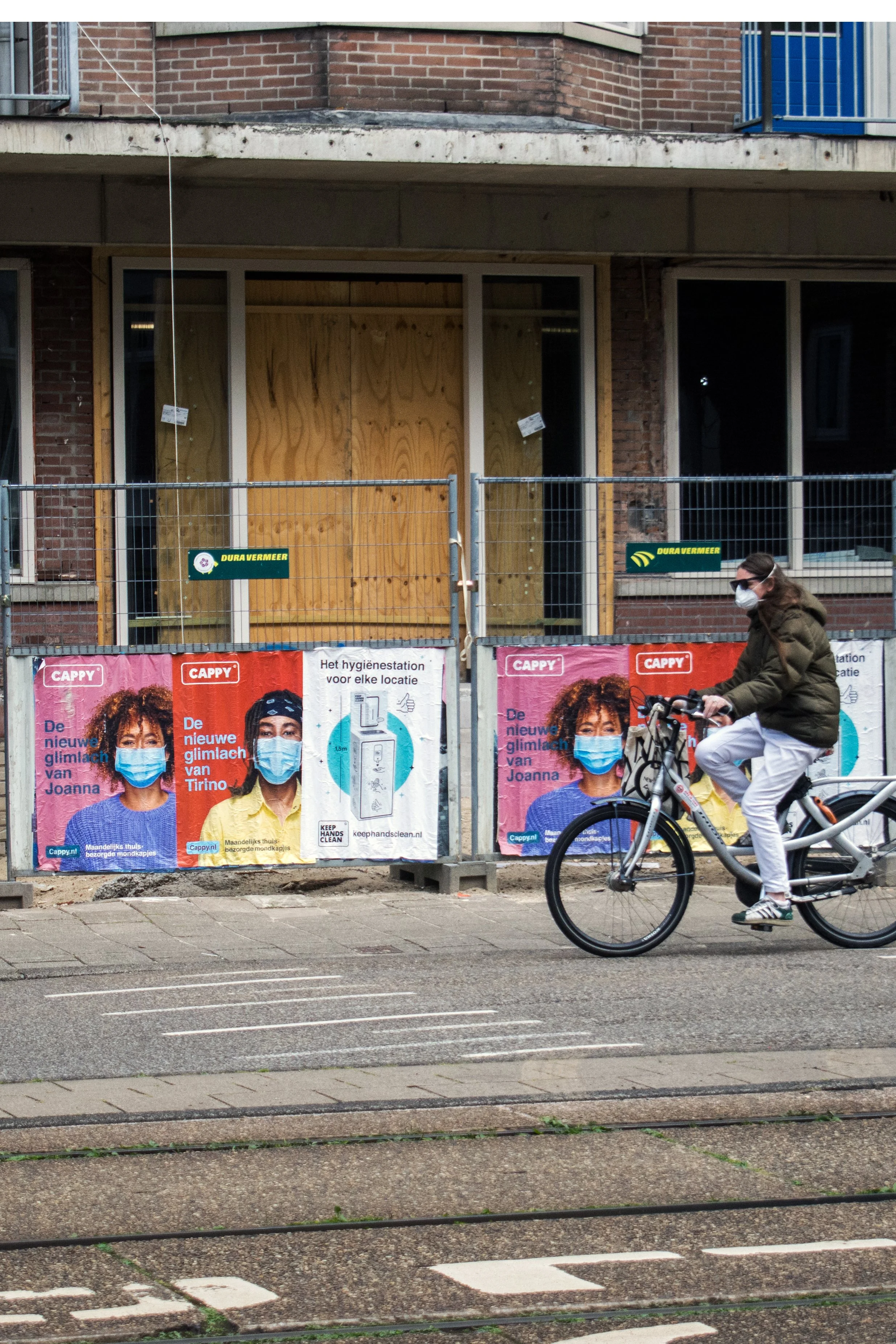 A woman riding a bicycle past a construction site with posters of women wearing face masks. The site has boarded-up windows and signs indicating construction work.
