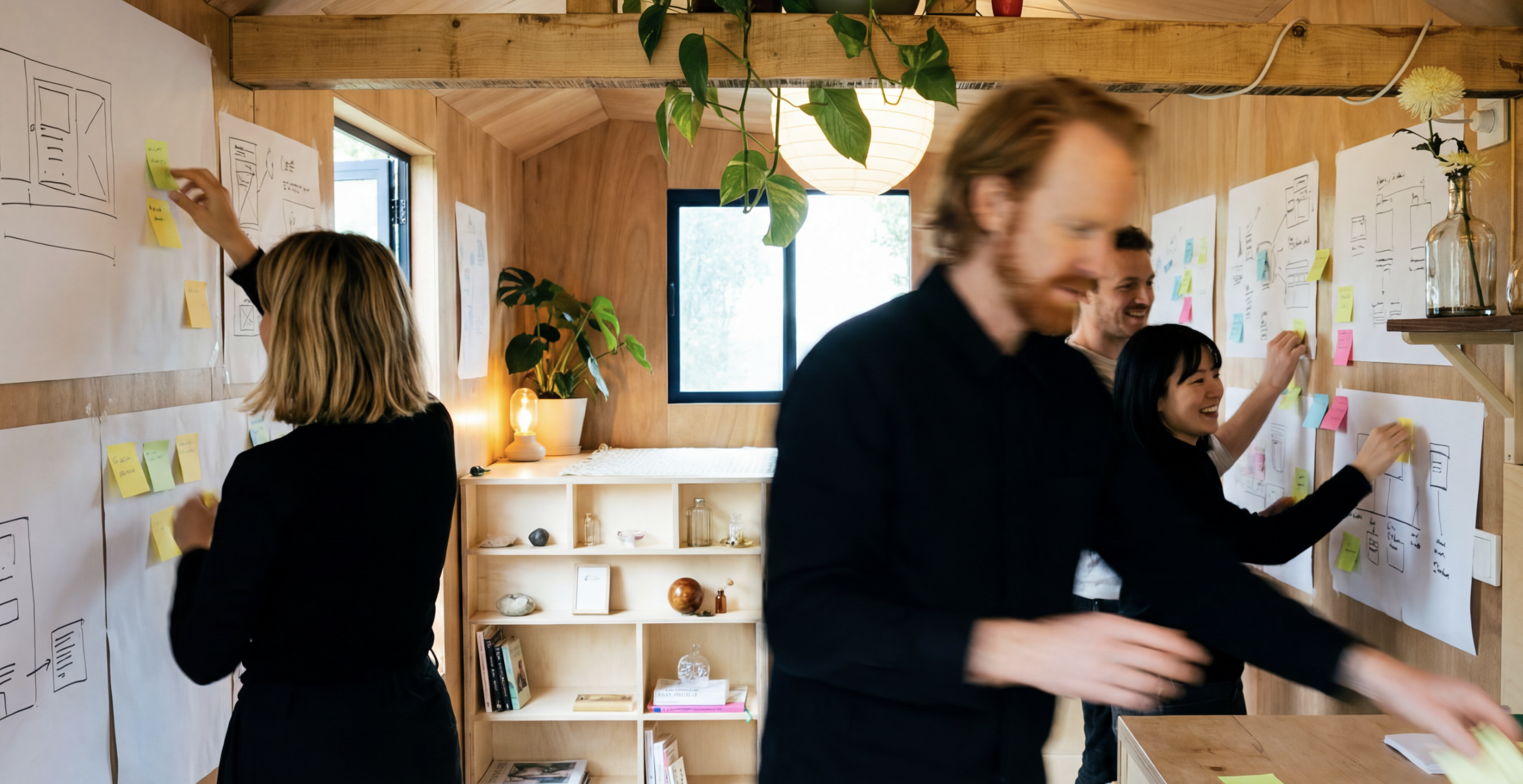 Group of four people collaborating on a project using sketches and sticky notes on papers taped to a wooden wall in a well-lit room with a window, plants, shelves, and decorative items.