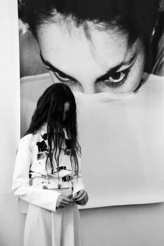 A woman with long hair standing in front of a large black-and-white portrait of a person's face with intense eyes, partially covering their mouth with a fabric.