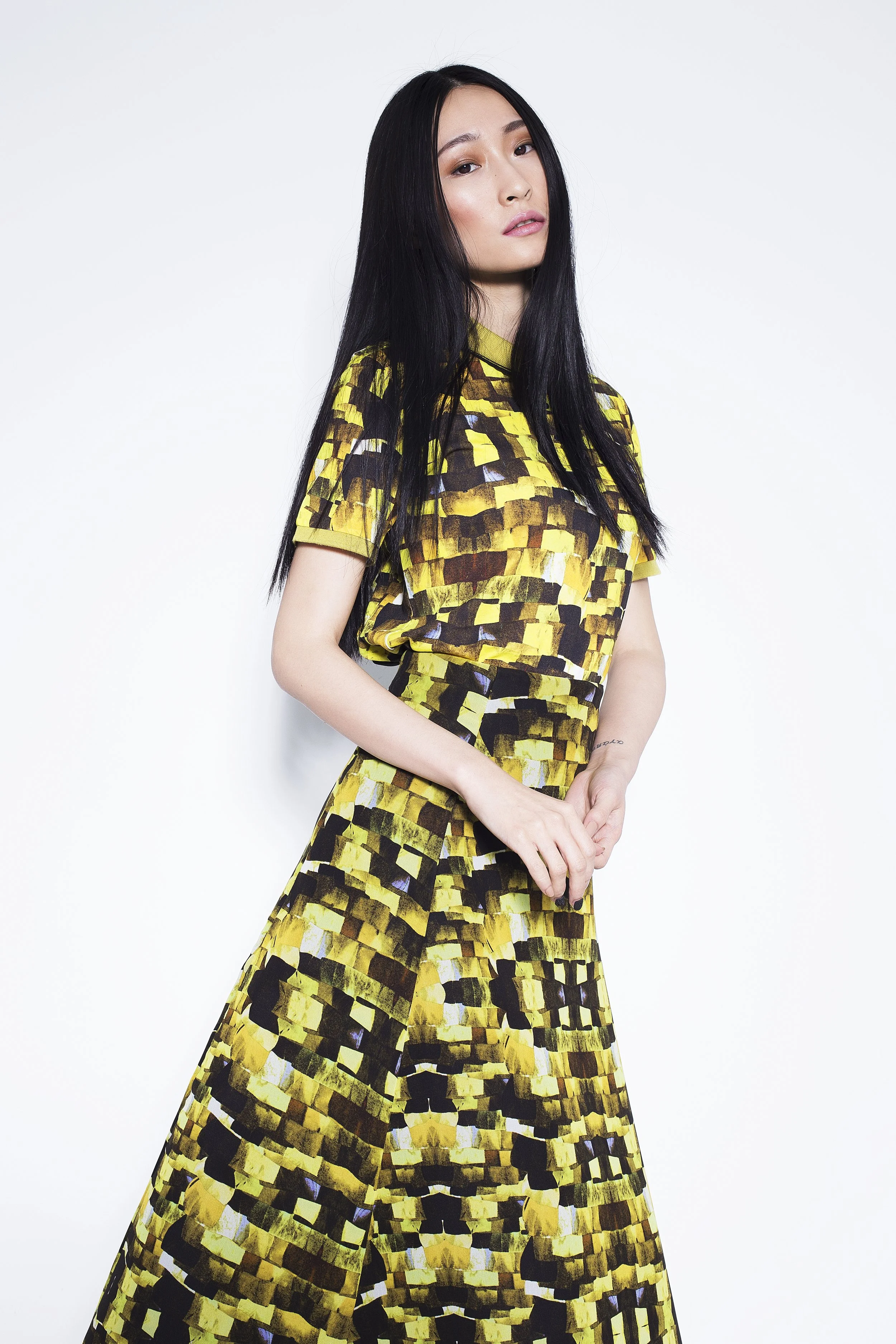A woman with long black hair wearing a yellow and black patterned dress standing against a plain white background.