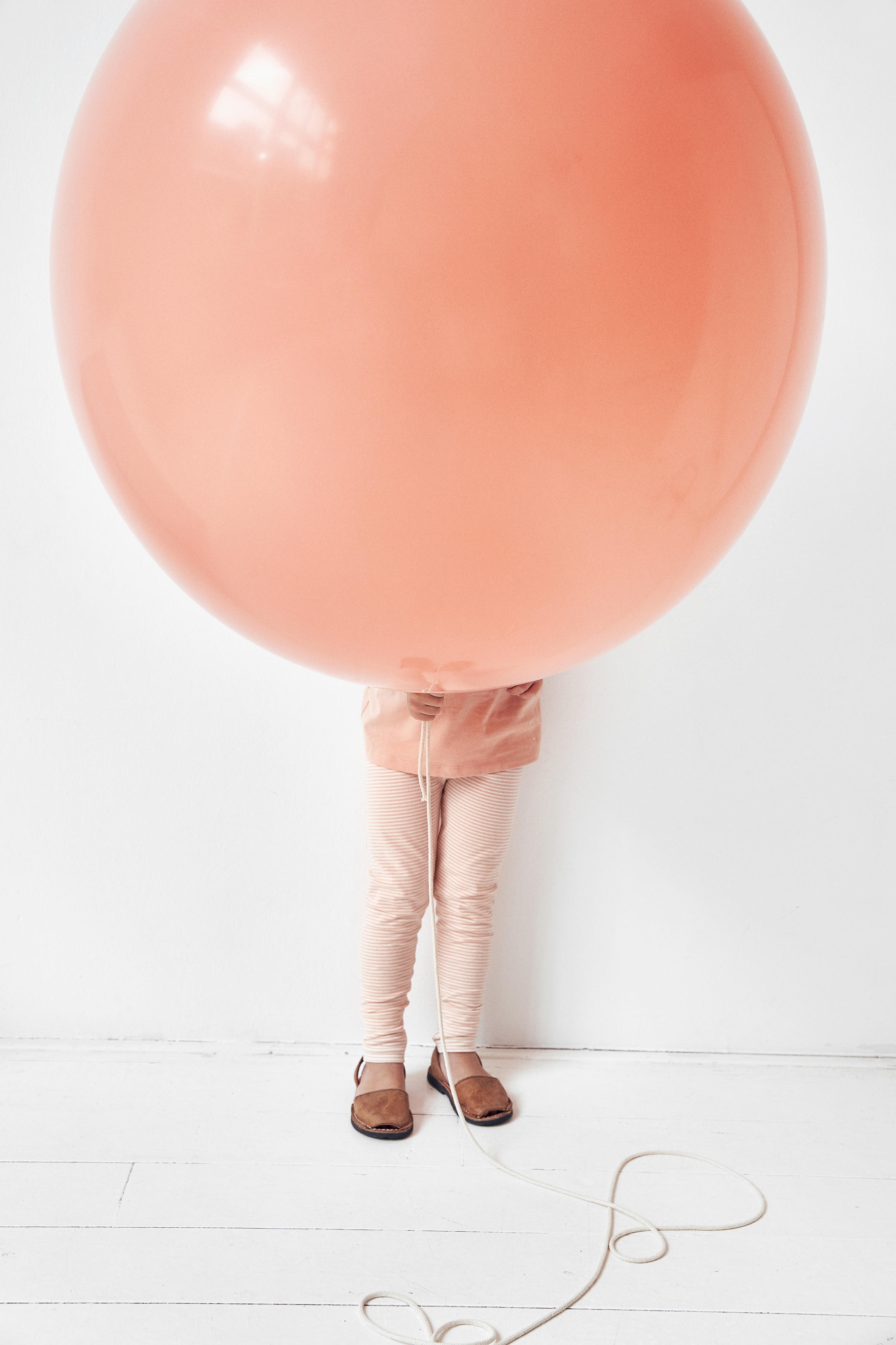 Person holding large pink balloon that covers their upper body and head, standing against a white wall and white floor, wearing beige shoes, striped pants, and a light-colored top.
