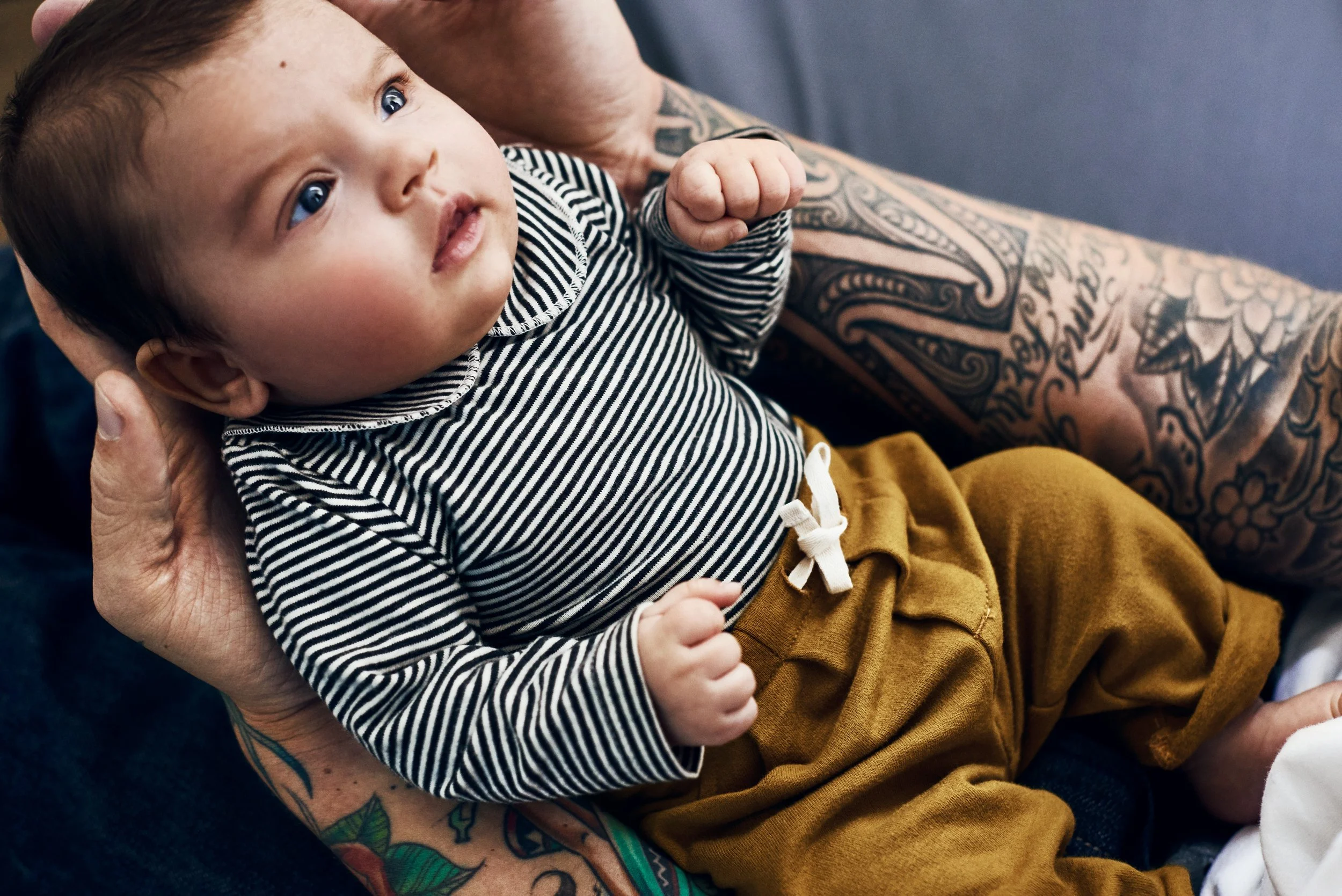 A baby with light skin and dark hair, wearing a black-and-white striped shirt and mustard-colored pants, is sitting on an adult's lap. The adult has visible tattoos on their arm and hand, and is holding the baby gently.