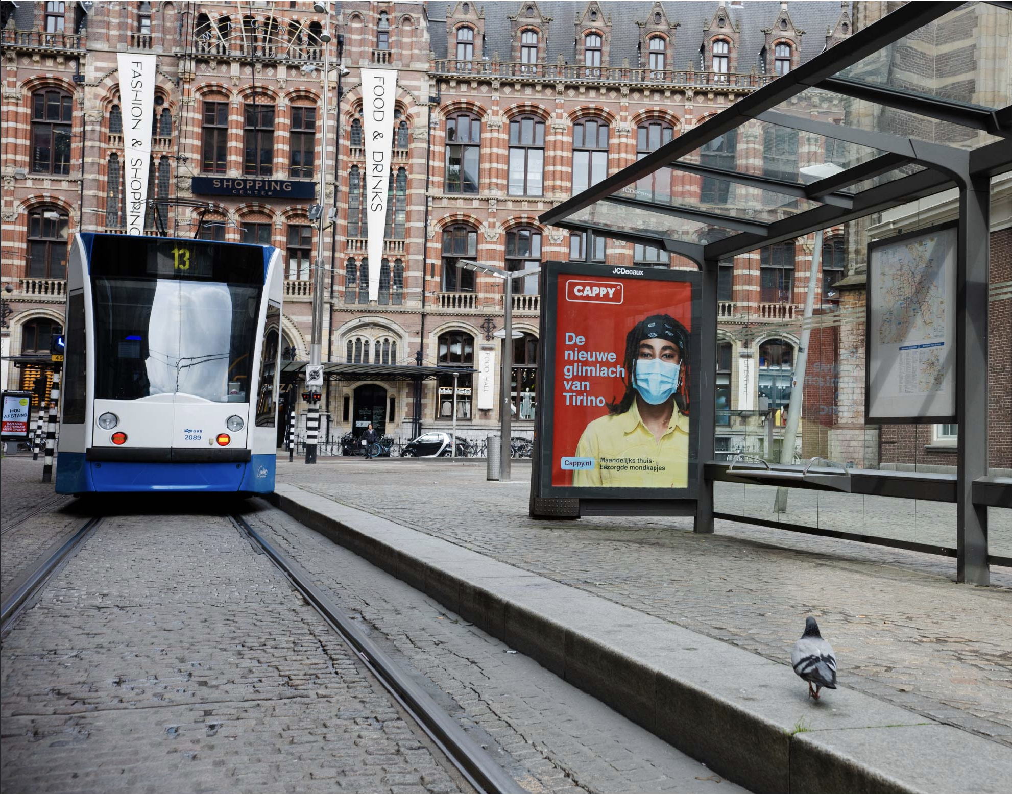 A street scene with a blue and white tram number 13 stopped at a tram stop. There is a bus shelter with an advertisement featuring a person wearing a face mask. In the background, a historic brick building with advertisements and a café sign is visible. A pigeon walks on the cobblestone sidewalk near the tram tracks.