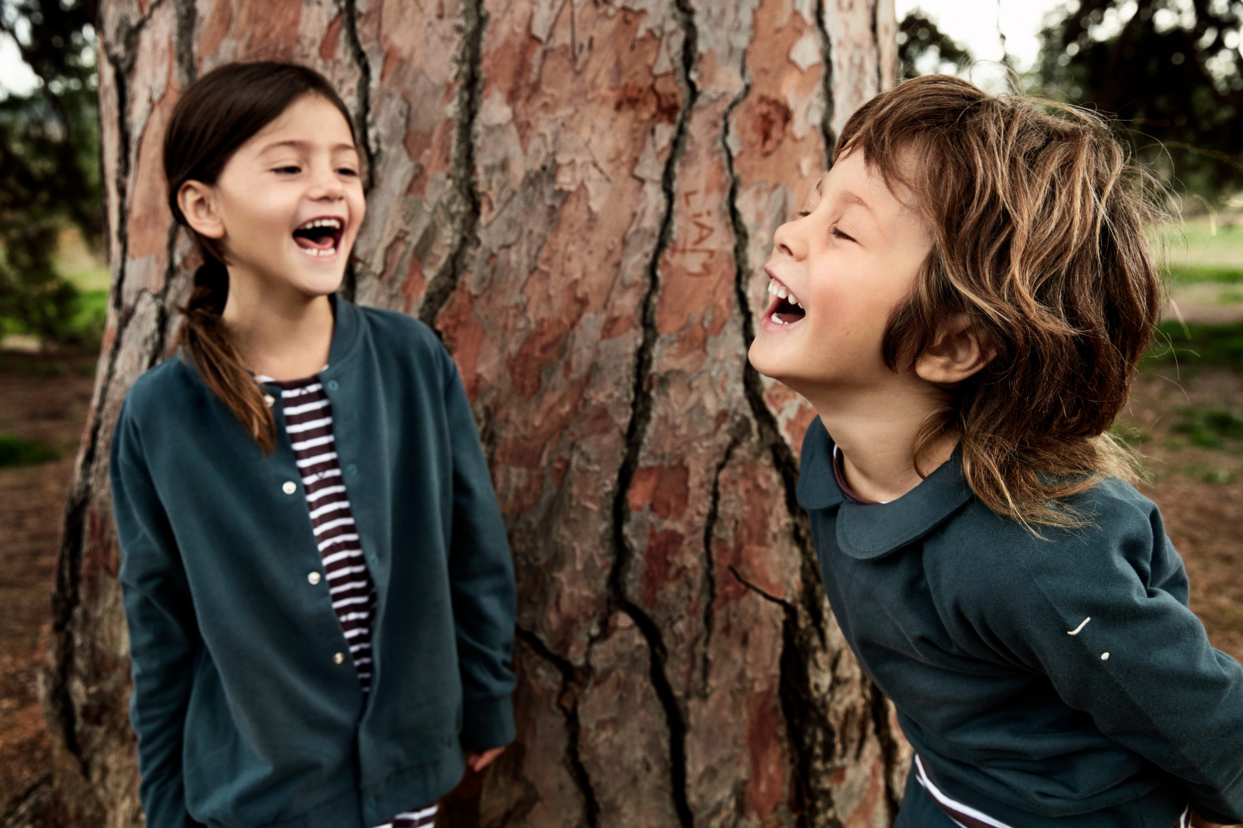 Two children, a girl with braided brown hair and a boy with curly brown hair, laughing and playing outdoors near a large tree with textured bark.