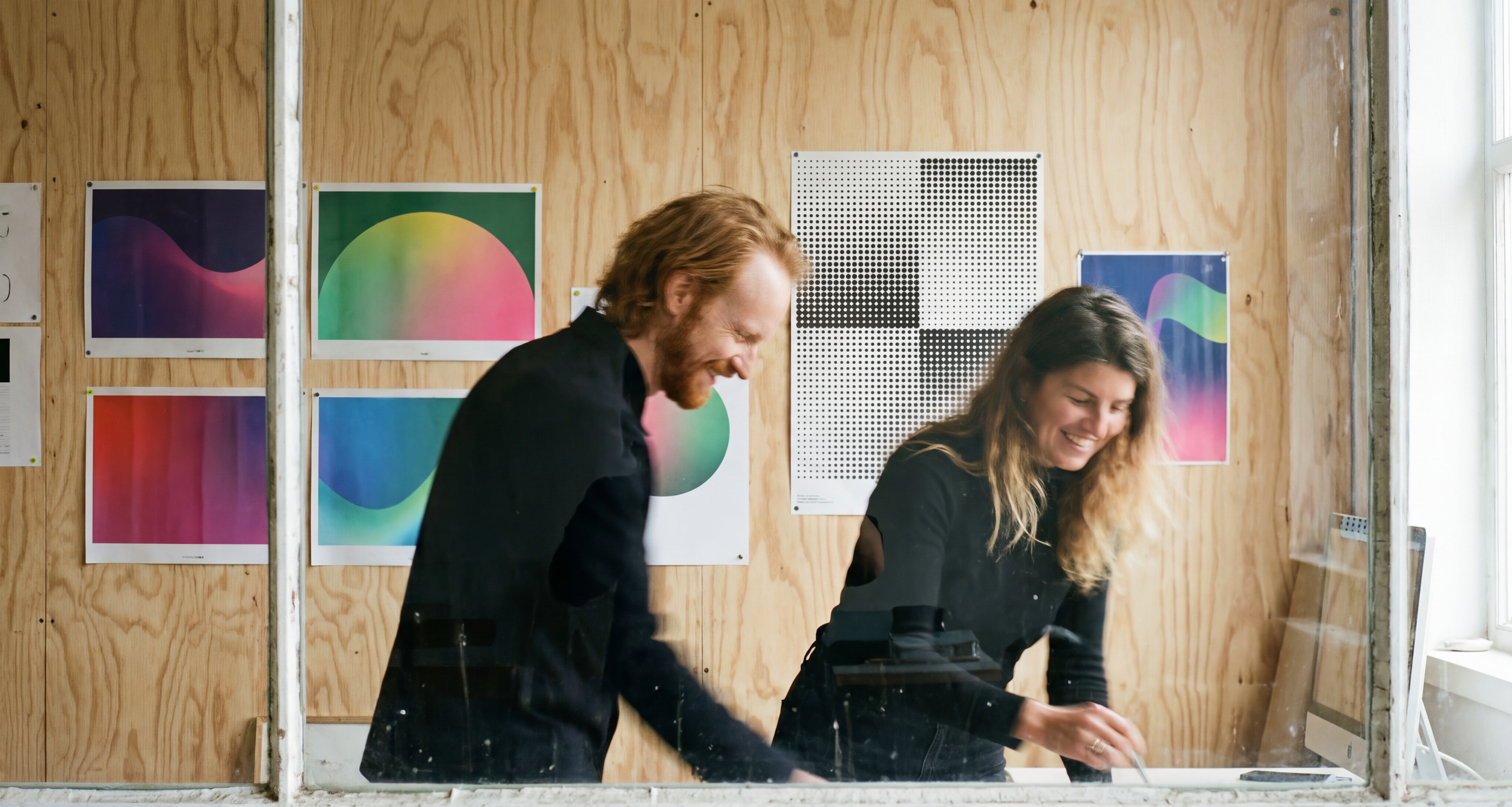 Two smiling women working together behind a glass window, with colorful abstract artwork on the wooden wall behind them.