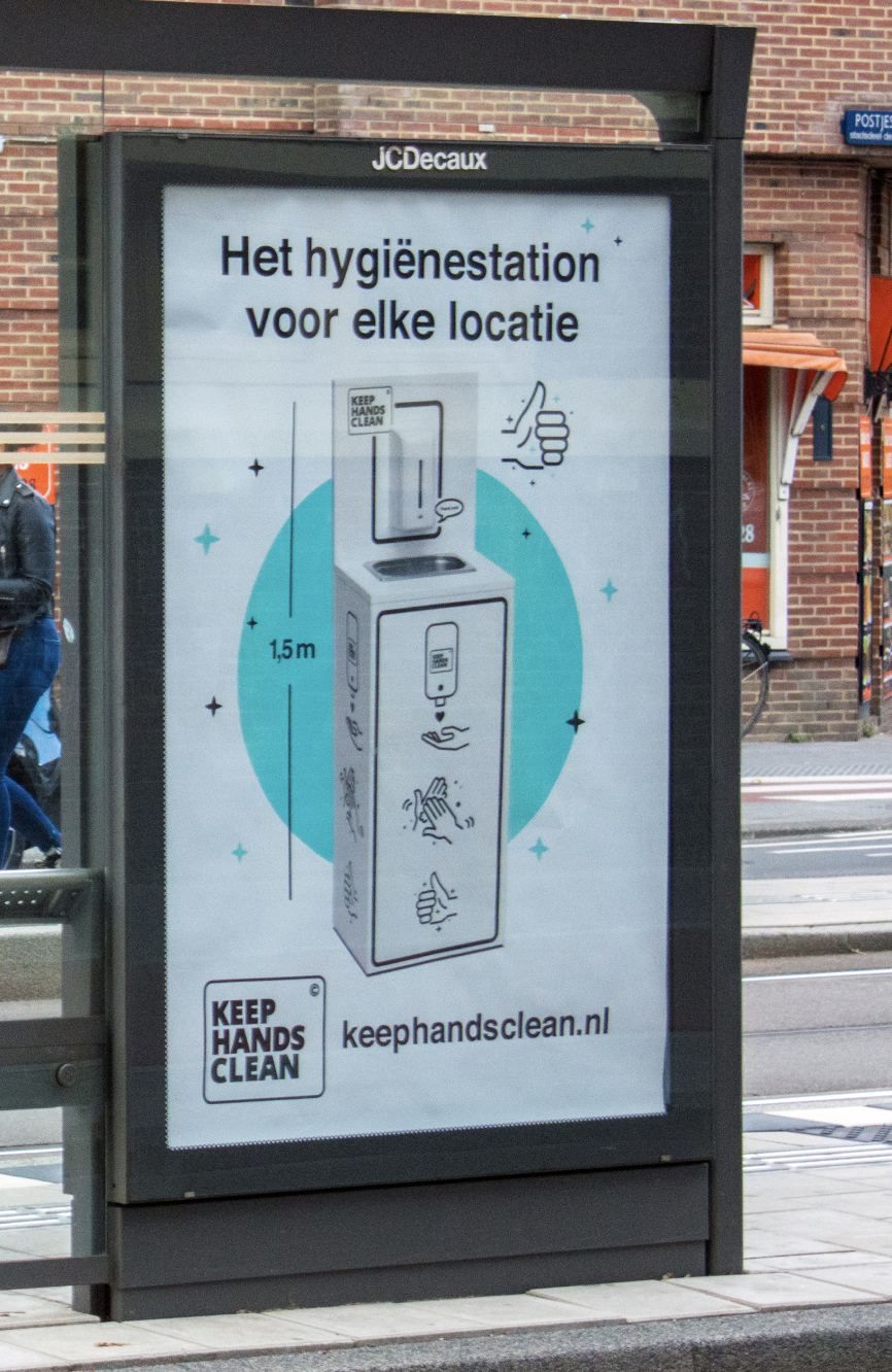A public hand sanitizing station at a bus stop with the sign in Dutch that says "The hygiene station for every location". It features a tall hand sanitizer dispenser, with step-by-step icons showing how to use it, a height of 1.5 meters, a message to keep hands clean, and a website kehehandsclean.nl.
