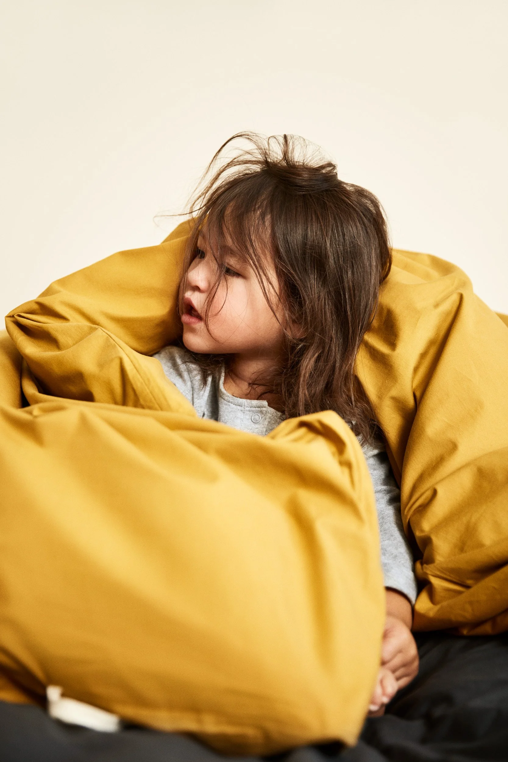 A young girl with brown, slightly messy hair looking to the left while leaning on a large yellow cushion or blanket.