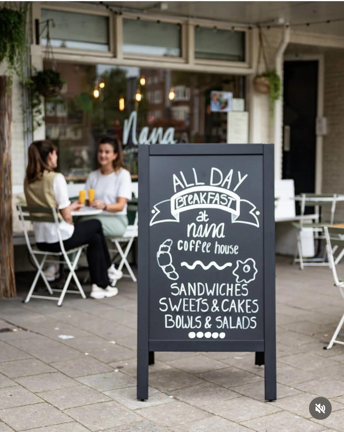 A chalkboard sign outside a cafe advertising all-day breakfast, sandwiches, sweets, cakes, bowls, and salads, with two women sitting at an outdoor table in the background, drinking orange juice.