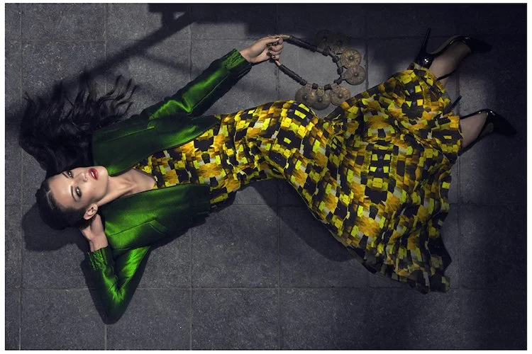 Woman lying on a dark floor, wearing a green jacket and a yellow patterned dress, holding a chain with circular links.