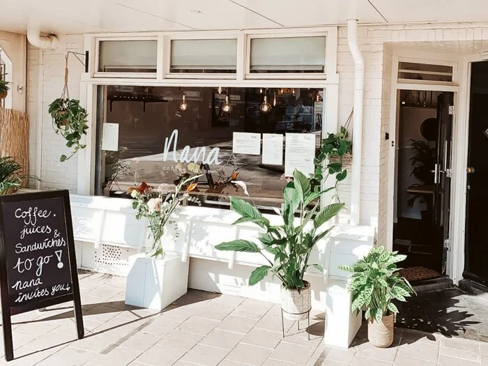 Exterior of a cozy coffee shop with the name 'Nana' on the window. Potted plants and a blackboard sign advertising coffee, juices, and sandwiches sit outside on the sidewalk.