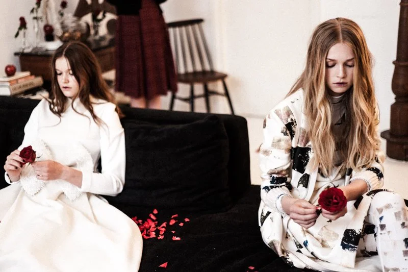 Two young women sitting on a black couch, each holding a red rose; one in a white dress with flower petals on her lap, the other in patterned pajamas, with a decorative wreath and scattered rose petals nearby.