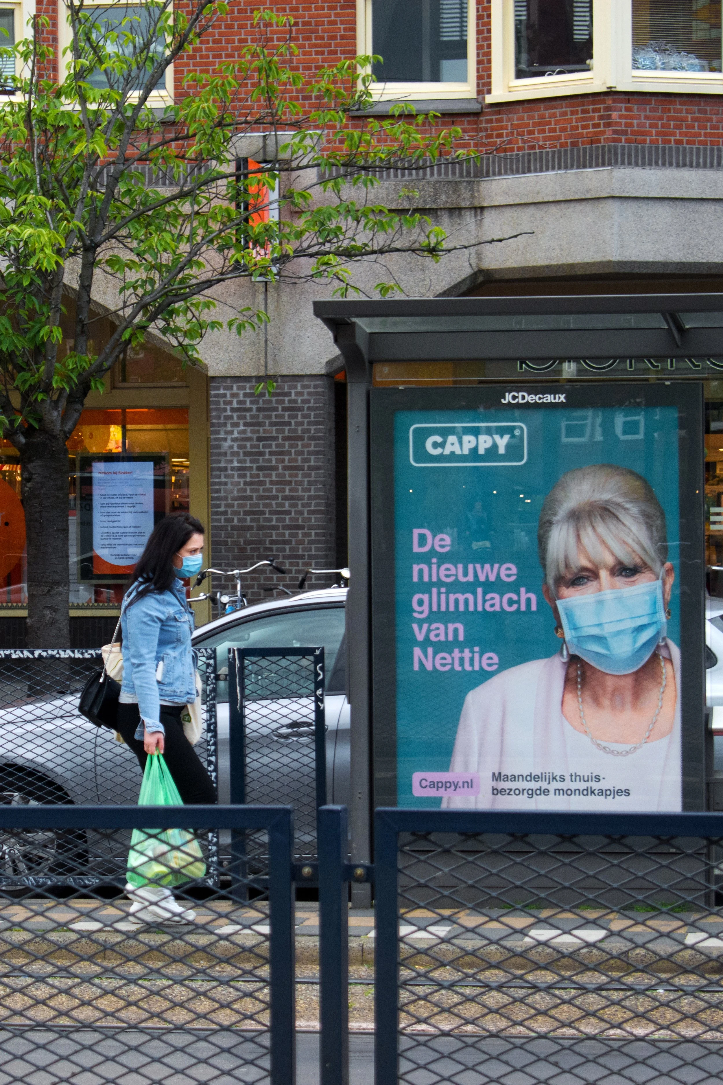 A woman wearing a face mask, jeans, and a denim jacket walking past a bus stop advertisement featuring a woman with gray hair wearing a face mask. The advertisement promotes a website called Cappy.nl and mentions monthly home-delivered face masks.