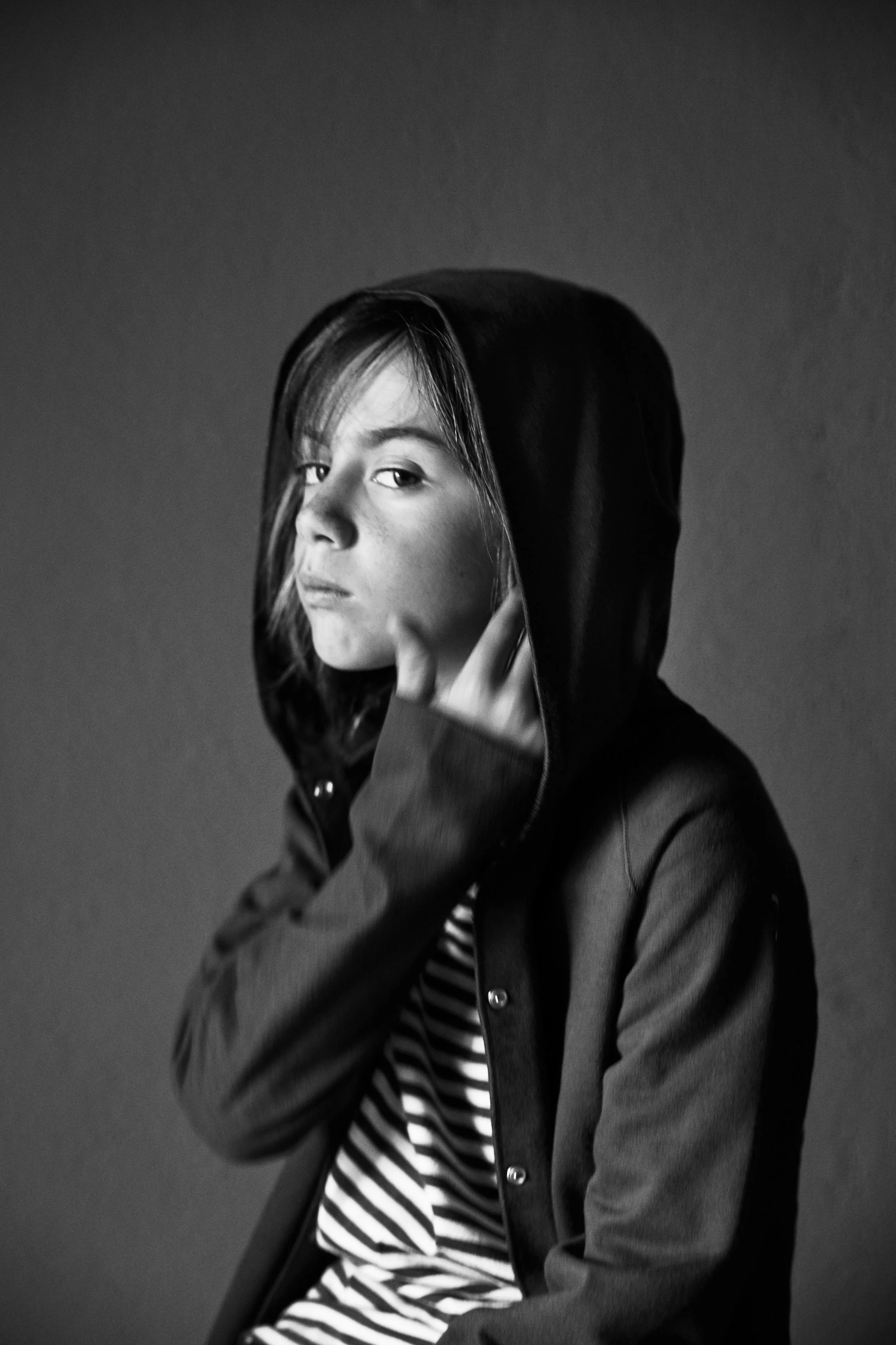 Black and white photo of a person wearing a hooded jacket, touching their face with one hand, and staring at the camera with a serious expression.