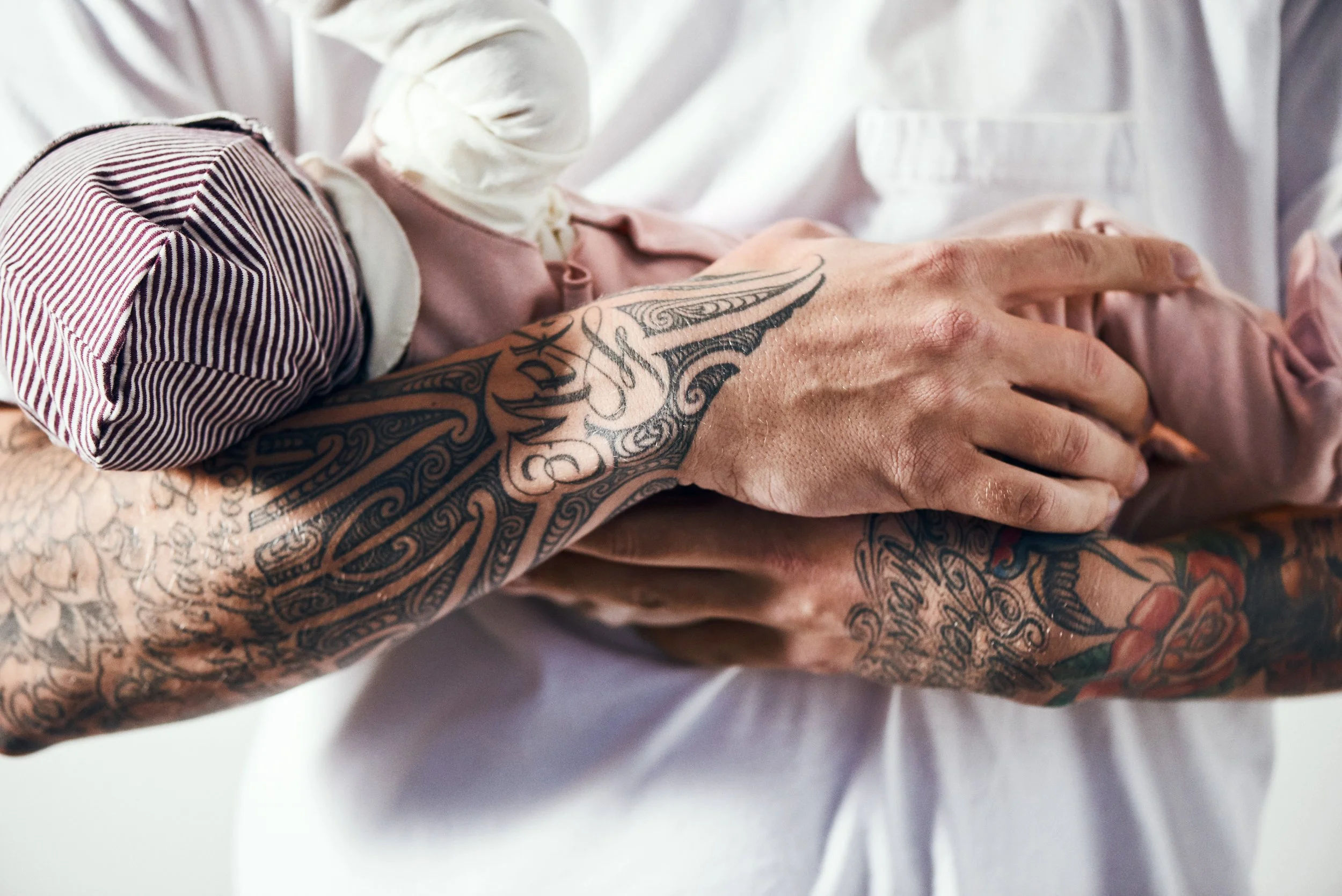 Close-up of two tattooed arms, one resting on the other's shoulder, with a person wearing a striped shirt and another in a white shirt.