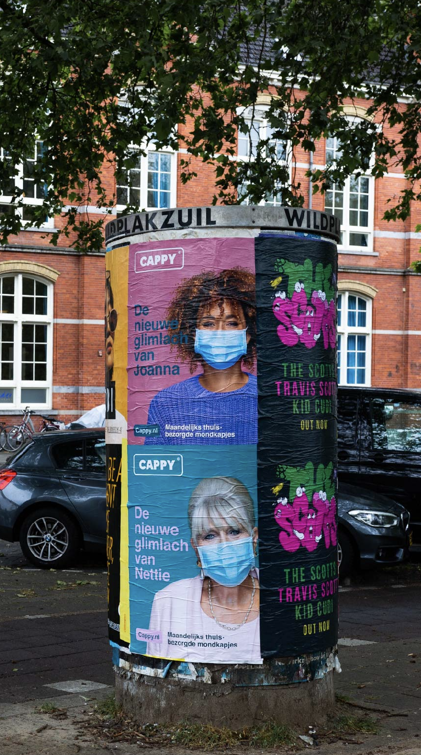 Two posters on a cylindrical pole in an urban setting, featuring women wearing face masks. The first poster shows a woman with curly hair and the second shows a woman with short blonde hair, both smiling with the text in Dutch. The background includes parked cars and a brick building with multiple windows.