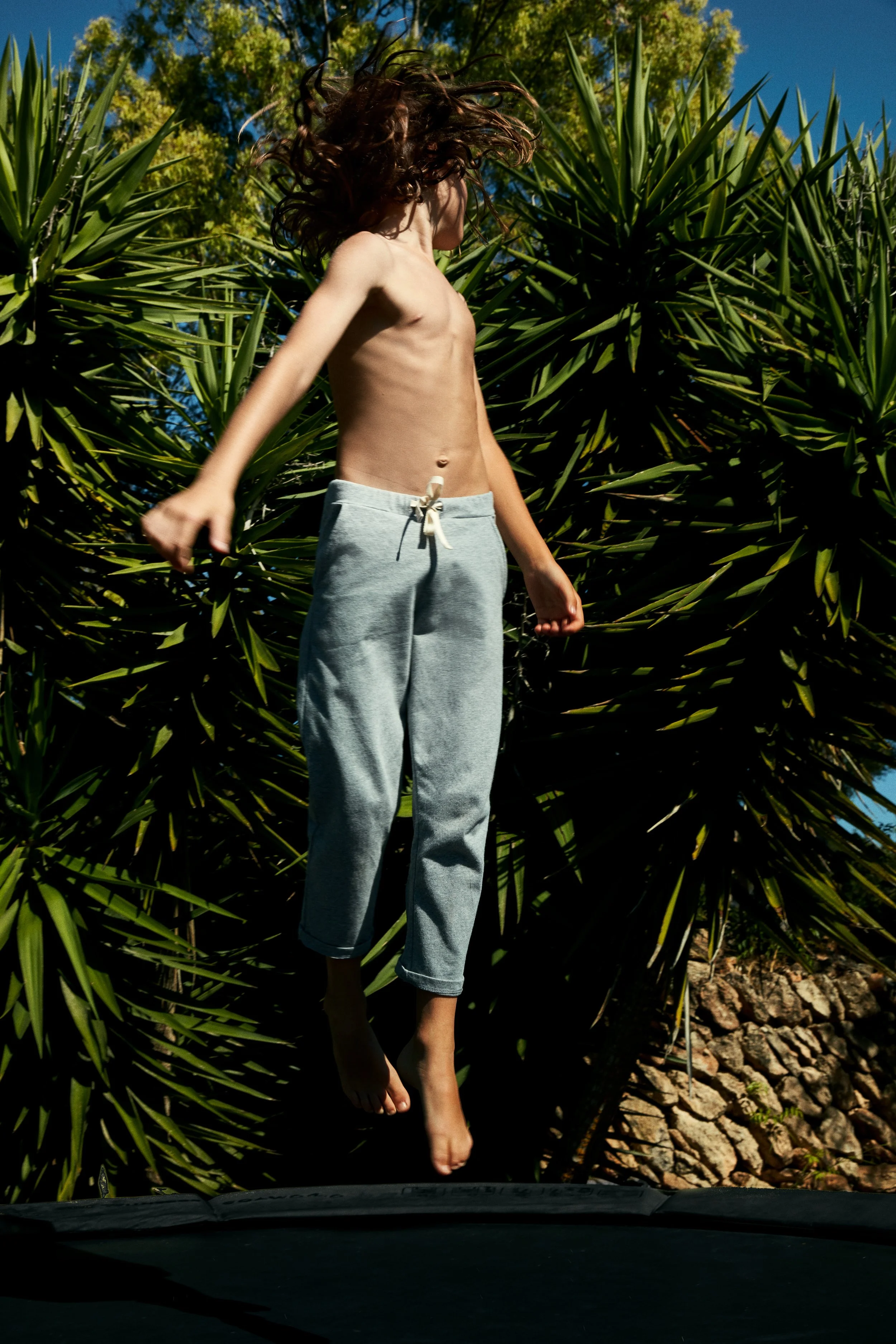 A shirtless boy with long hair jumping on a trampoline outdoors during daytime, with green foliage and trees in the background.