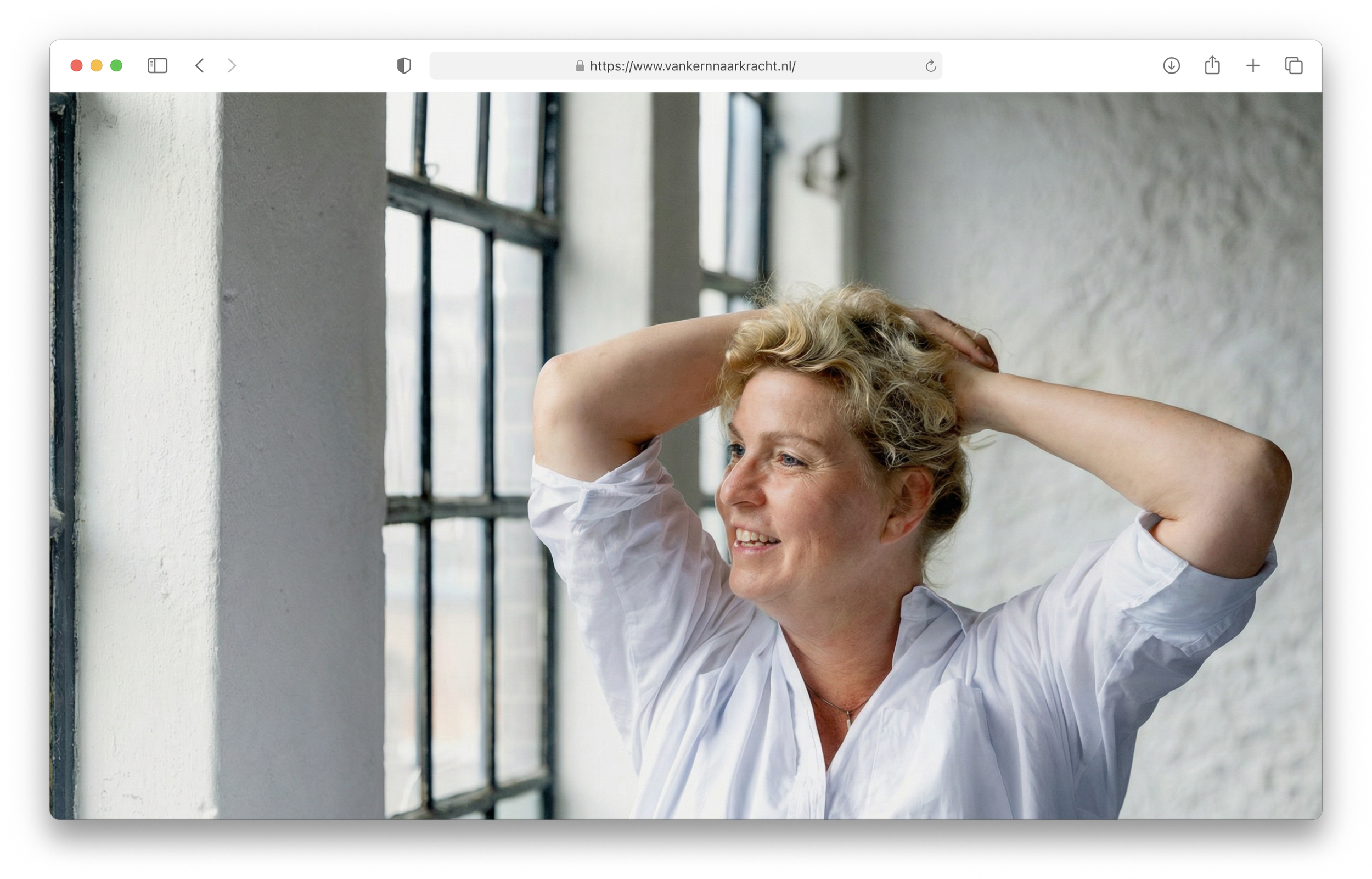 A woman with short curly gray hair smiling, wearing a white shirt, standing near a window with metal framing, with hands behind her head, in a bright room.